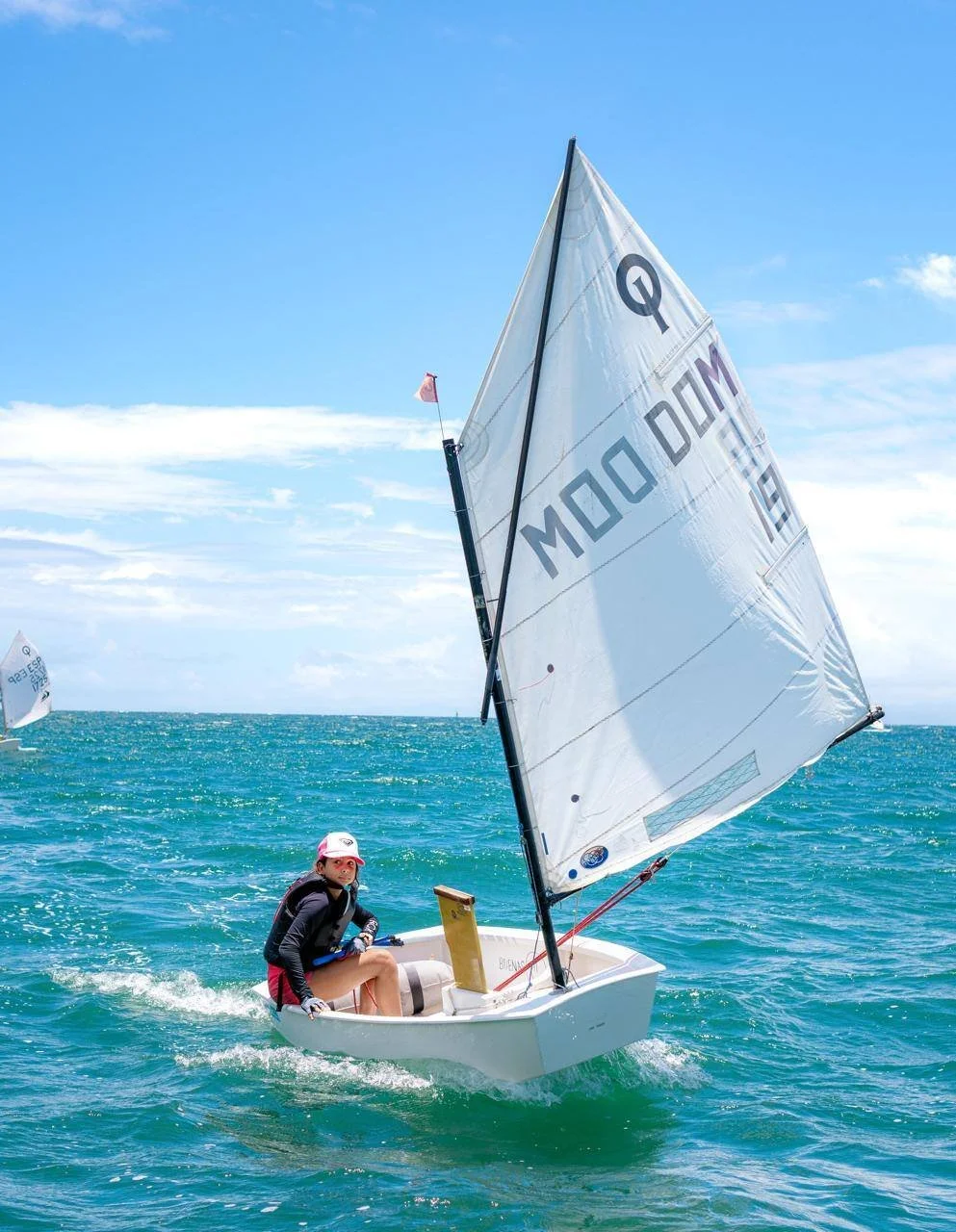 A person sailing a small sailboat on open water with a blue sky overhead. The sailboat has a large white sail with markings, and the person is wearing a life jacket, hat, and casual clothing.