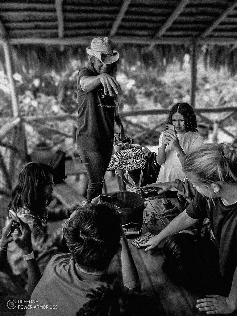 Group of women and children gathered around a table outdoors, with one woman standing and pointing, another woman taking a photo, and others engaging with items on the table in a rustic, open-air setting.