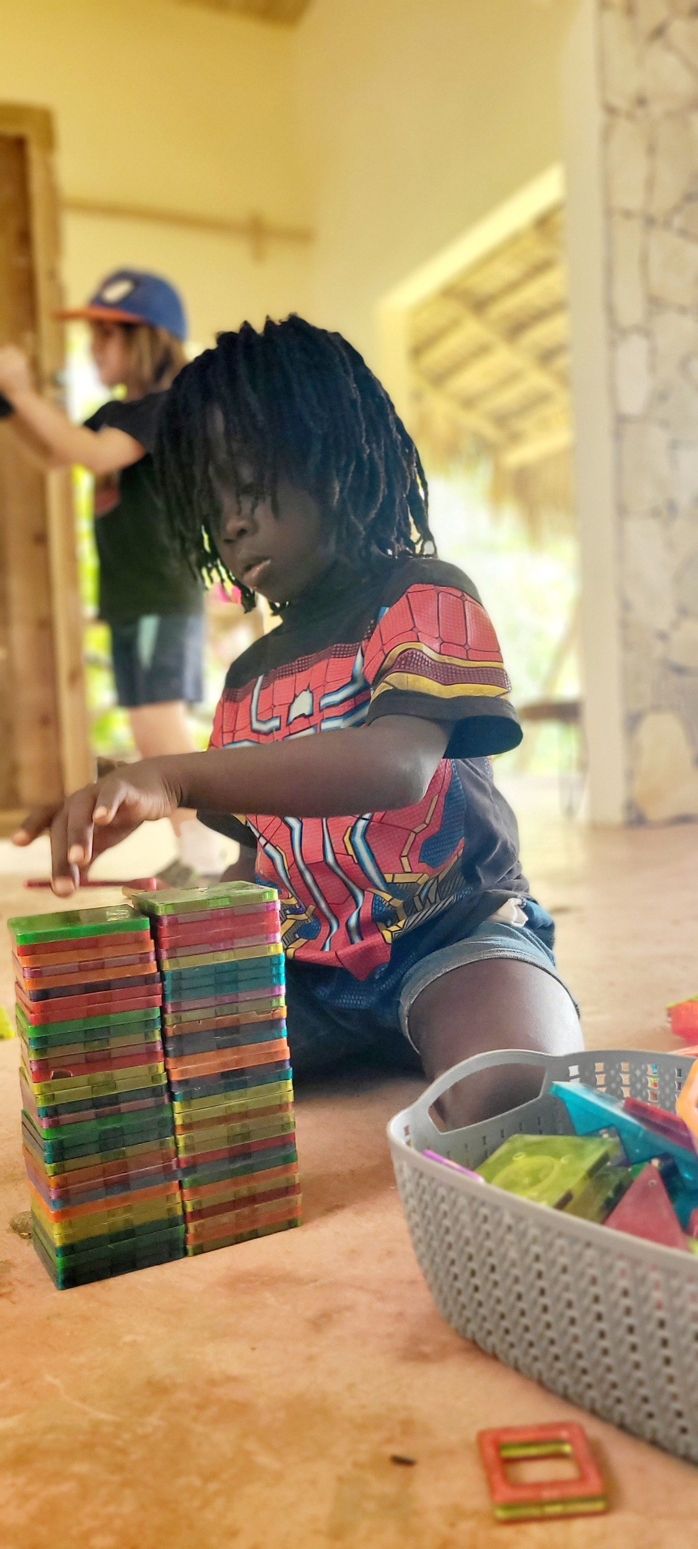 A young boy with dreadlocks playing with colorful connecting tiles on a wooden surface. Another child in a blue cap and black shirt is in the background.