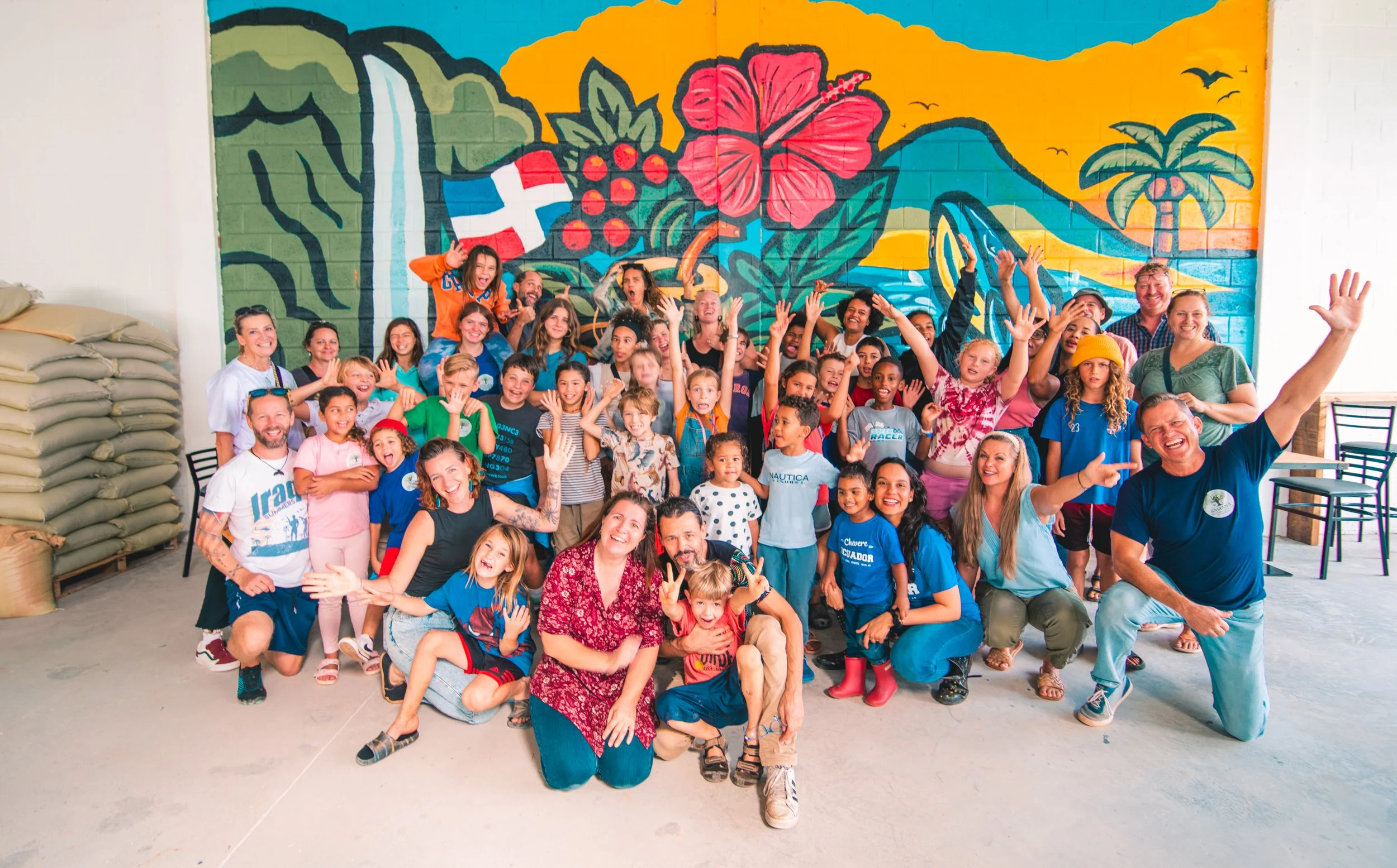 Group of children and adults posing happily in front of a colorful mural of a tropical scene with a volcano, palm trees, and a hibiscus flower