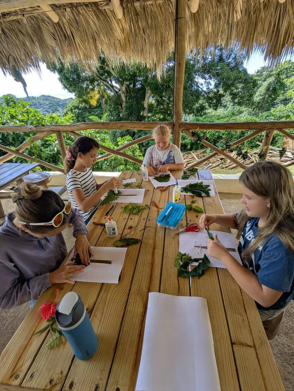 Four girls at a wooden table under a thatched roof, working with leaves and flowers, with trees and greenery in the background.