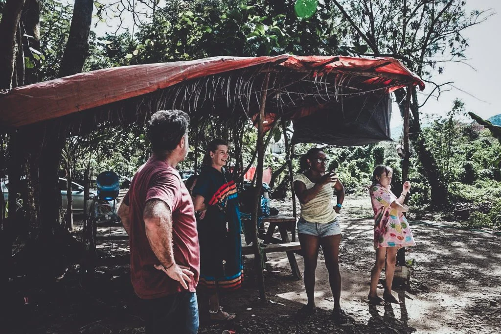 Group of five people standing and talking near a makeshift shelter in a wooded outdoor area, with trees and sunlight in the background.