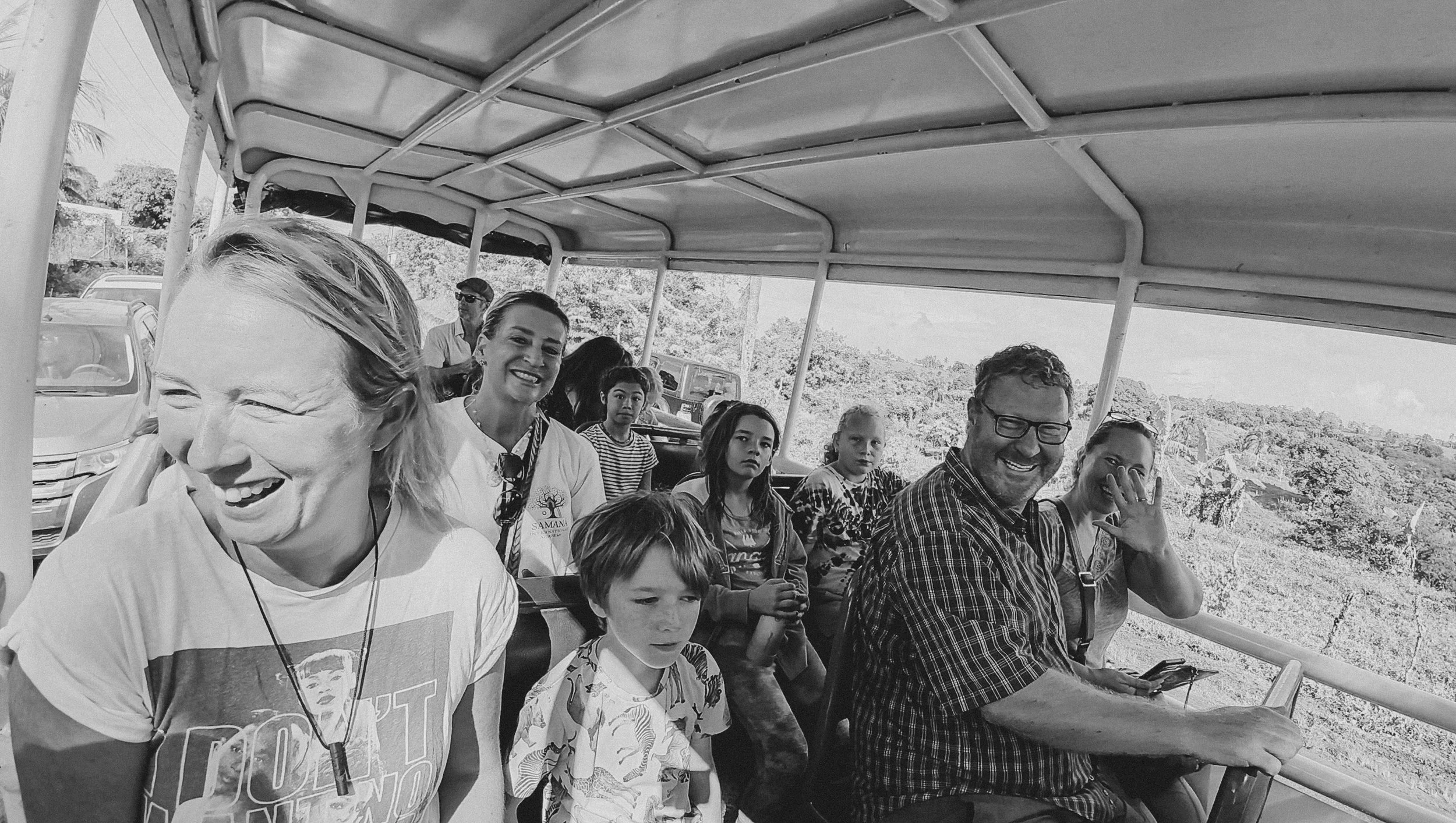 A group of smiling and laughing people riding a tram or bus, with some of the children looking serious, against a backdrop of trees and vehicles outside the window.