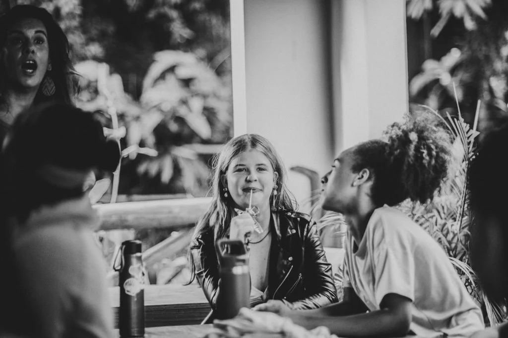Three women engaged in conversation at a table indoors, with a window showing greenery outside.
