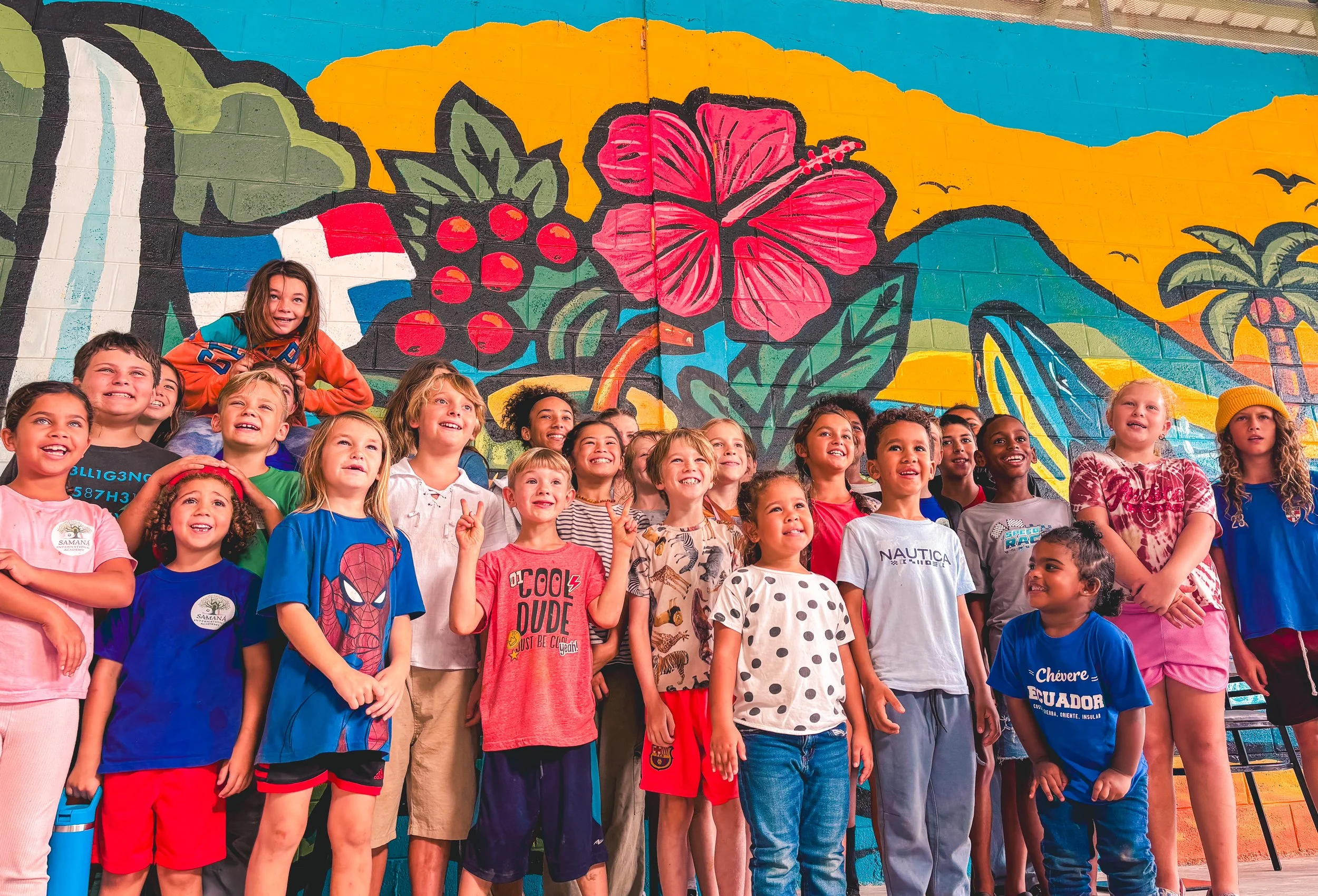 Children smiling and gathered in front of a colorful mural featuring tropical flowers, berries, and a palm tree.