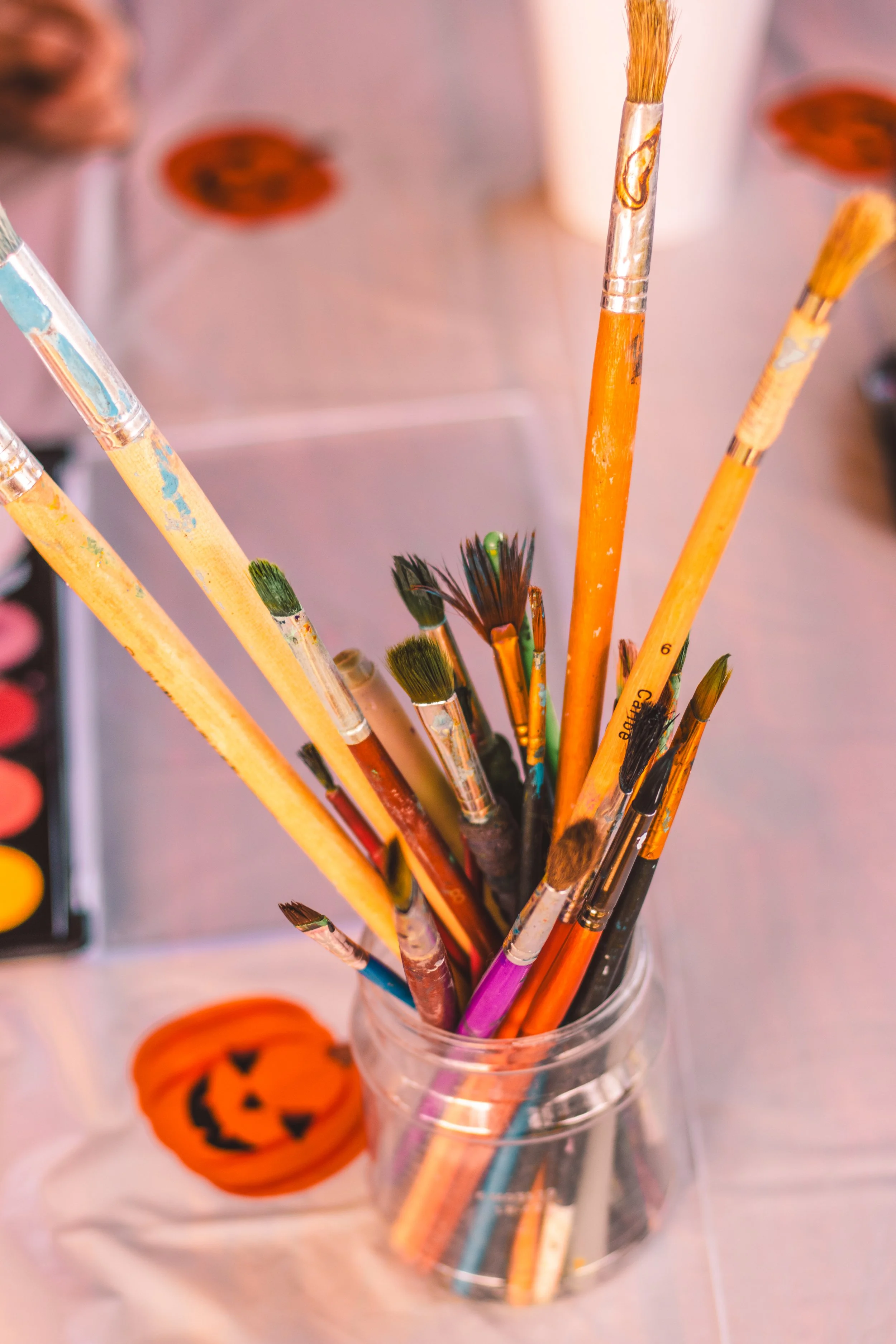 A collection of paintbrushes in a clear container with Halloween themed decorations in the background.