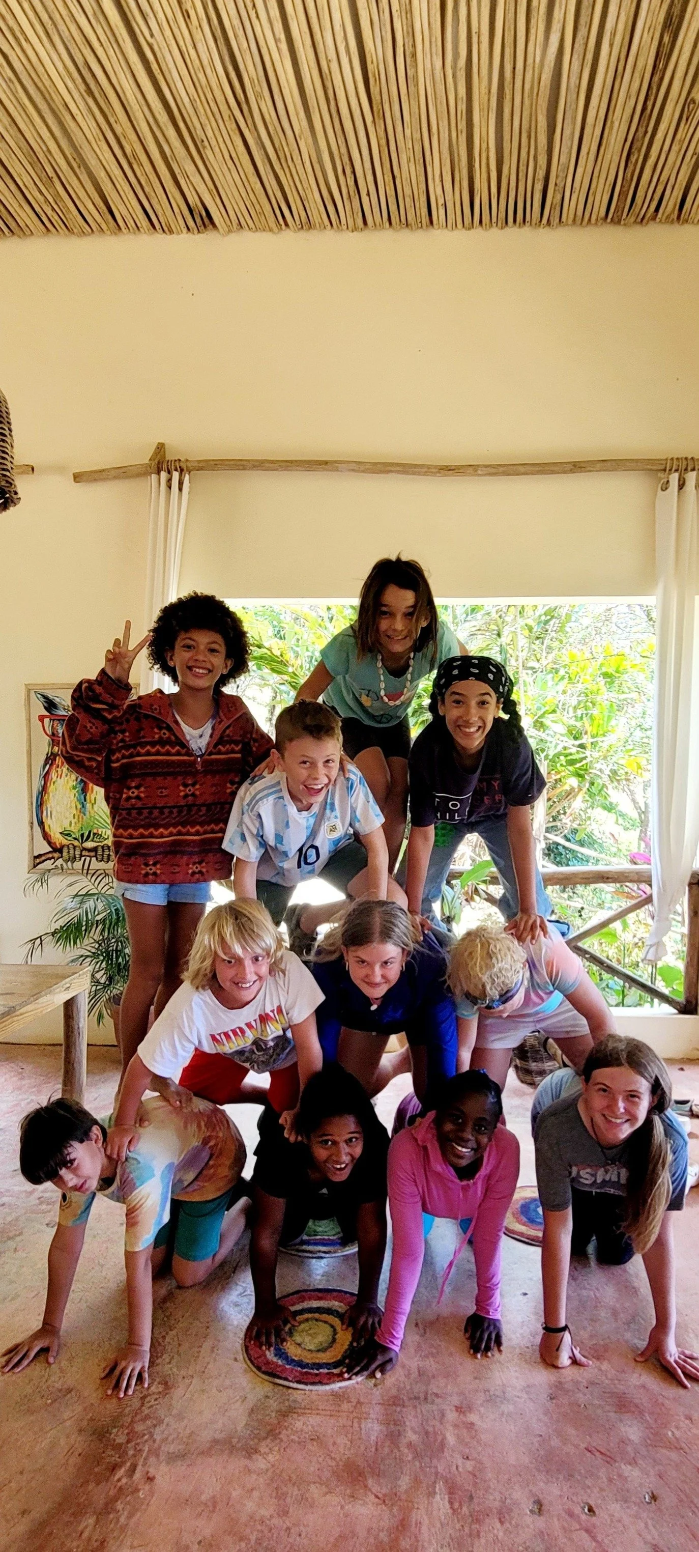 A group of children forming a human pyramid indoors near large window with green foliage outside, smiling and making peace signs.