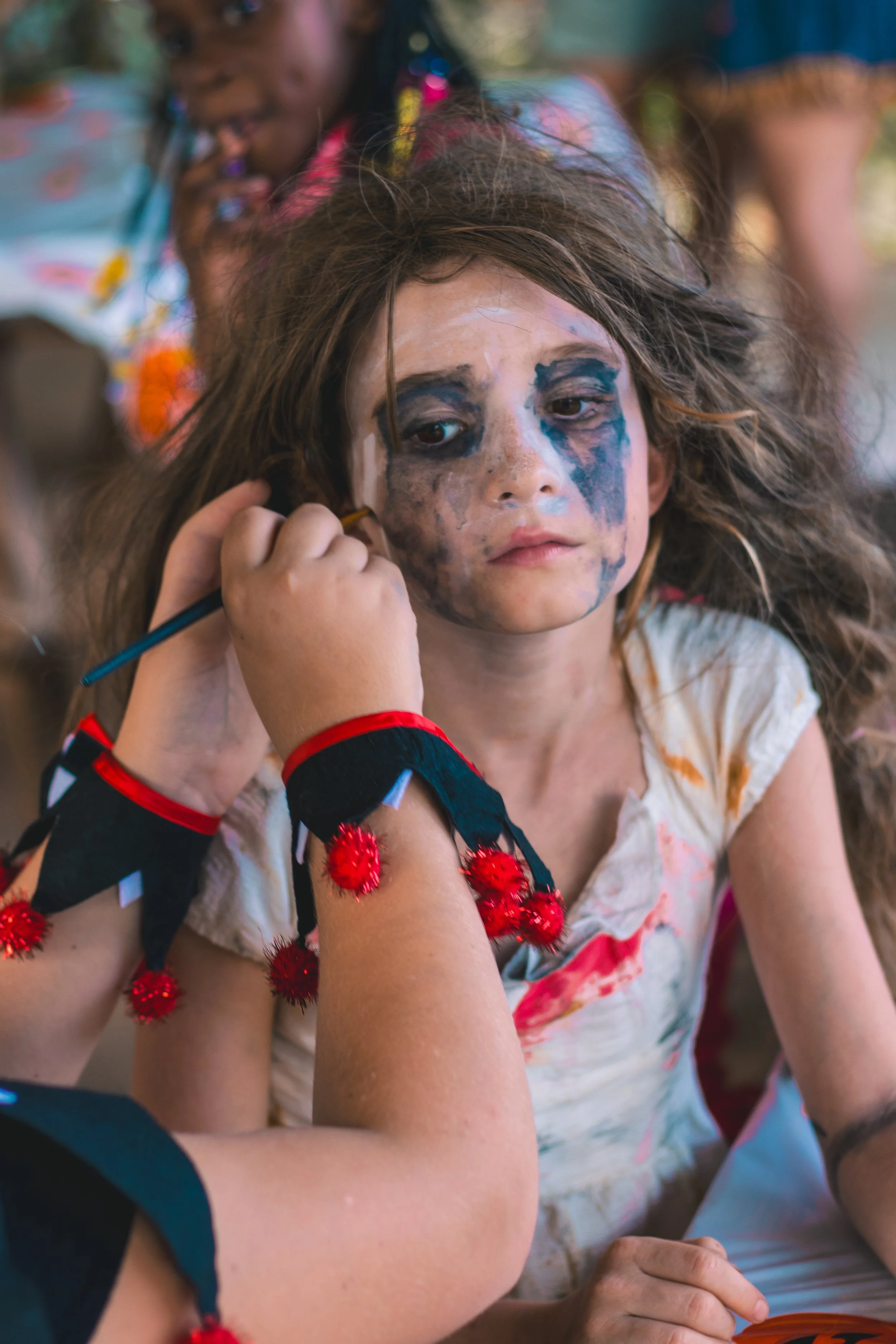 A young girl with face paint and a Halloween costume sitting at a table, with another child in the background. The girl has messy hair and a neutral expression.