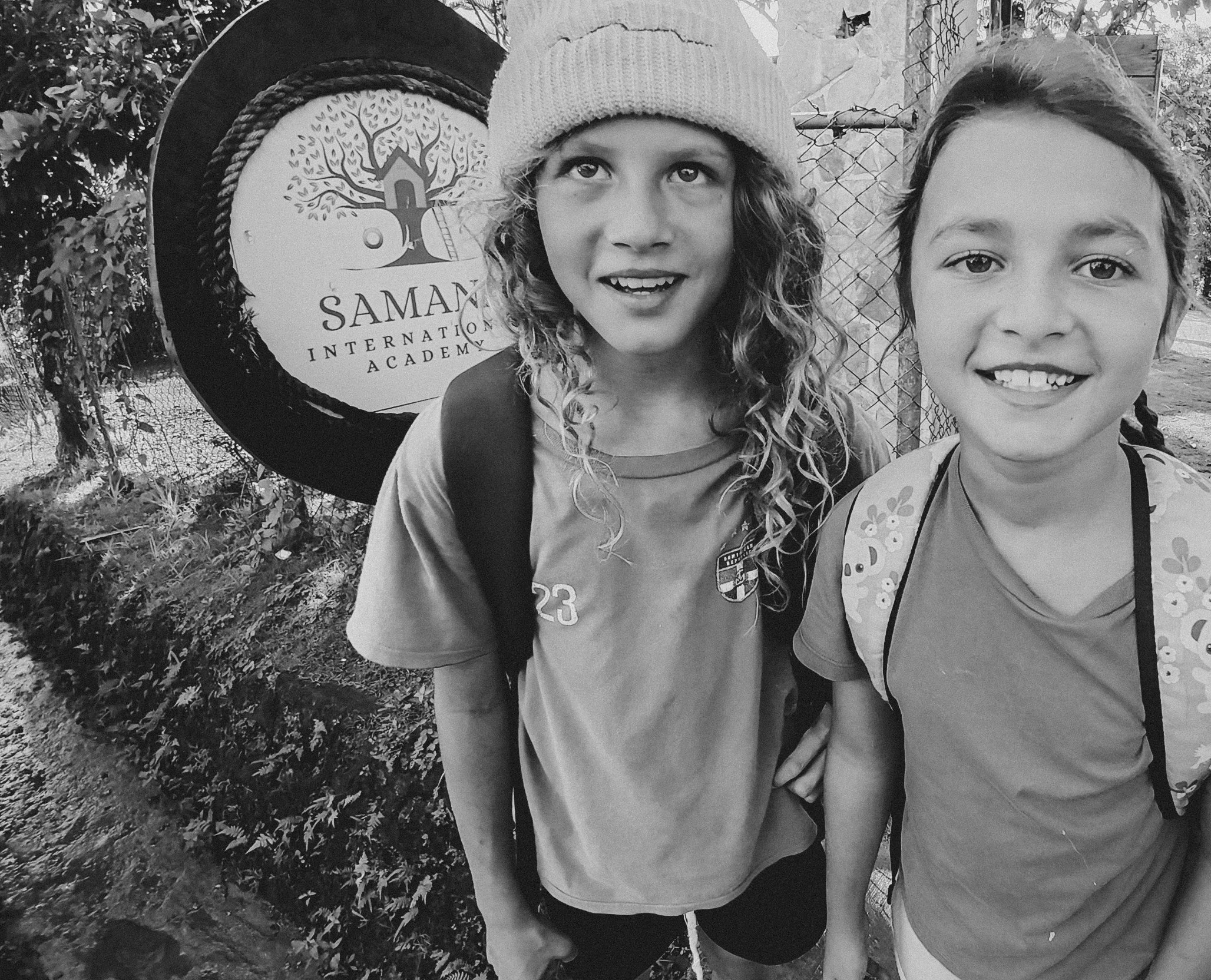 Two young girls standing outdoors, smiling for the camera, with a sign behind them that reads "Saman International Academy." The girl on the left has curly hair, wears a beanie and a backpack, while the girl on the right has straight hair, wears a pa