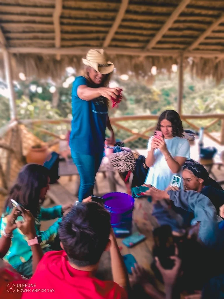 Group of people gathered around a woman standing on a wooden table, taking photos and videos with smartphones in a rustic outdoor setting.