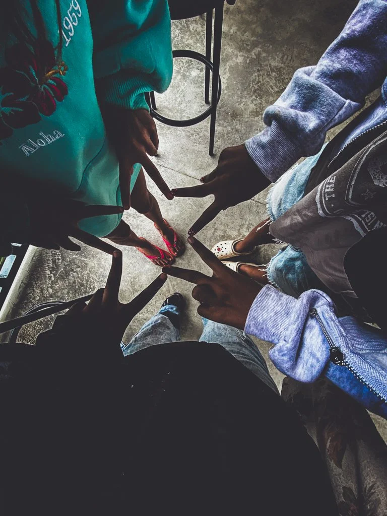 Six people standing in a circle, holding hands with their fingers interlaced, viewed from above. The floor is a gray concrete surface.