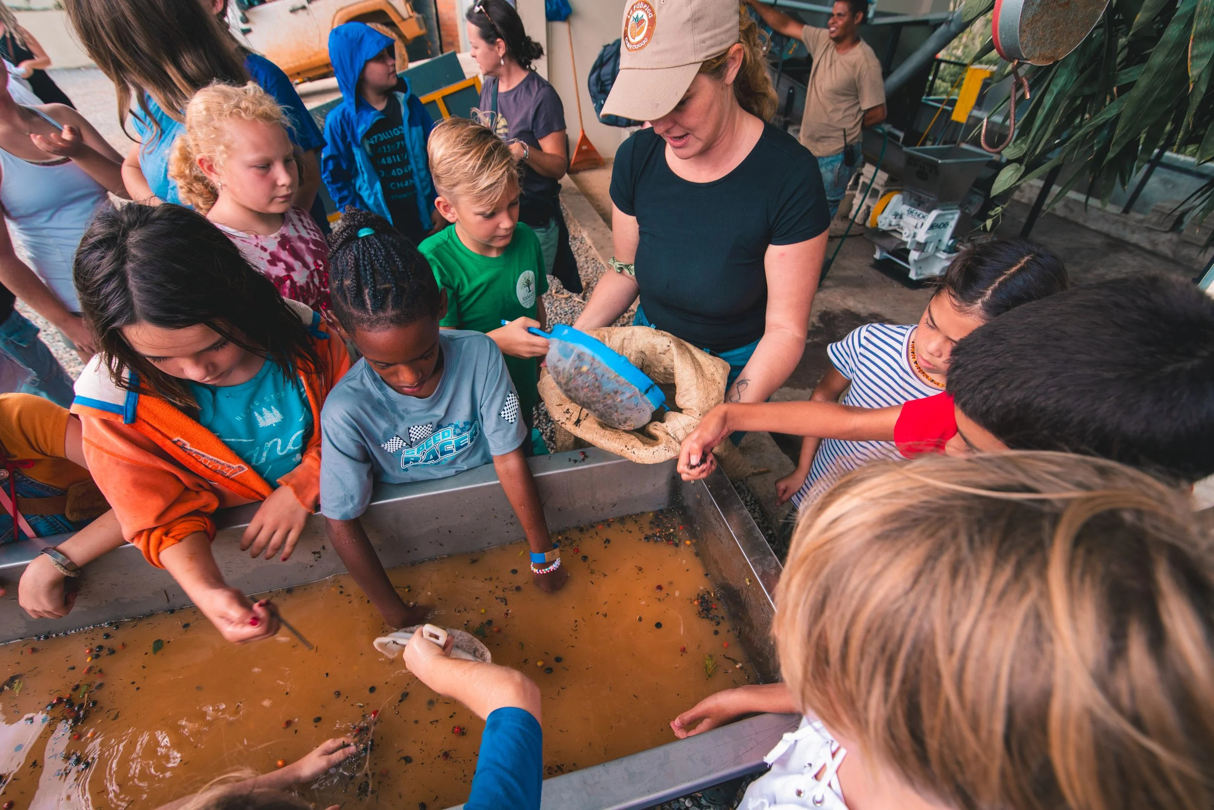 A group of children gathered around a water tank with brown muddy water, observing and reaching into the water while a woman in a black shirt and tan hat explains or demonstrates something.