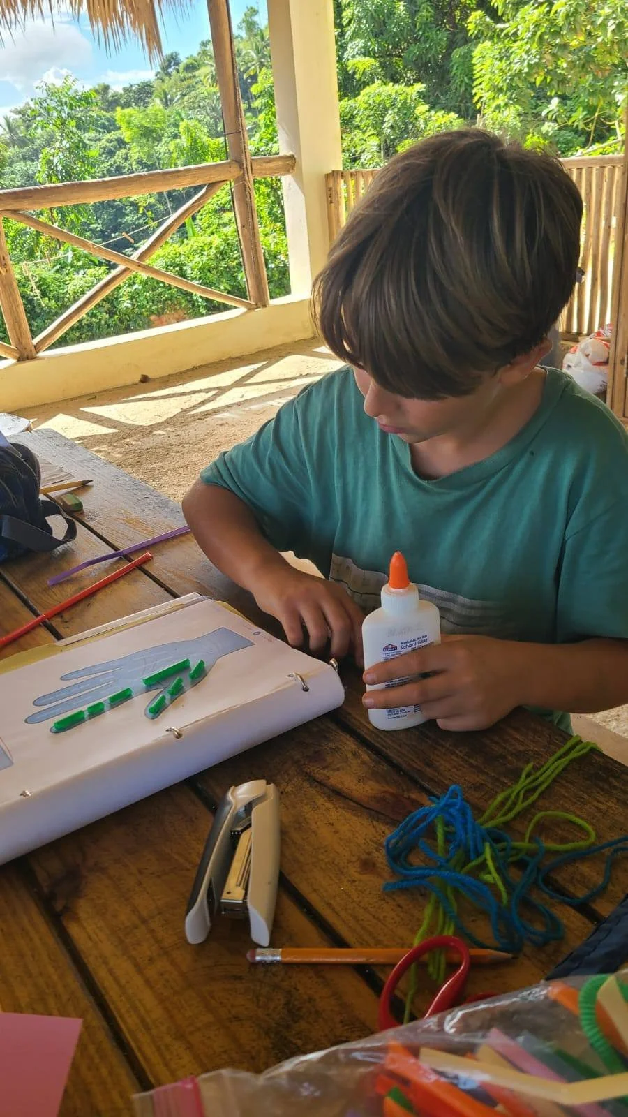 A boy sits at a wooden table outdoors, using glue to attach green foam pieces to a paper hand template, surrounded by colorful craft supplies, with lush green trees and a blue sky in the background.