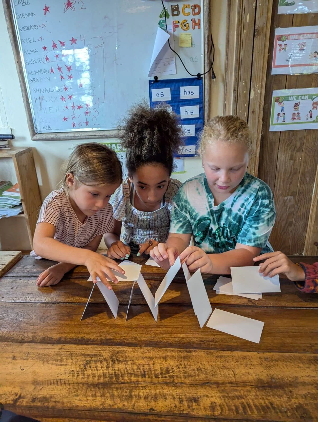 Three children working together at a wooden table, folding and assembling white paper cards in a classroom.