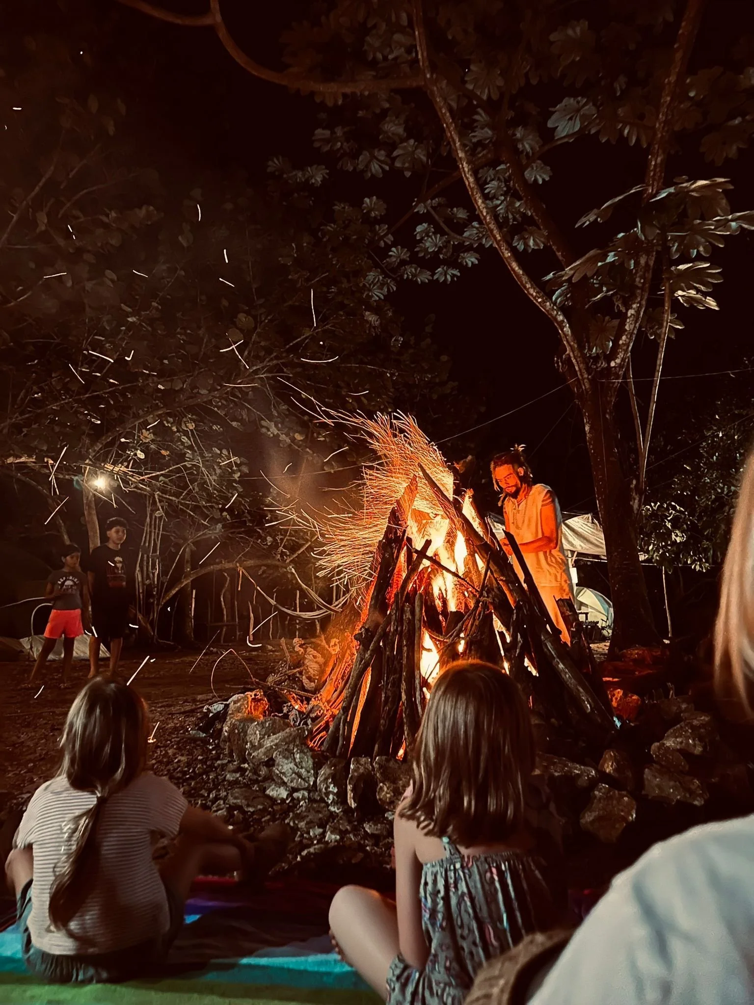 People sitting around a campfire at night, with trees overhead and children watching the fire.