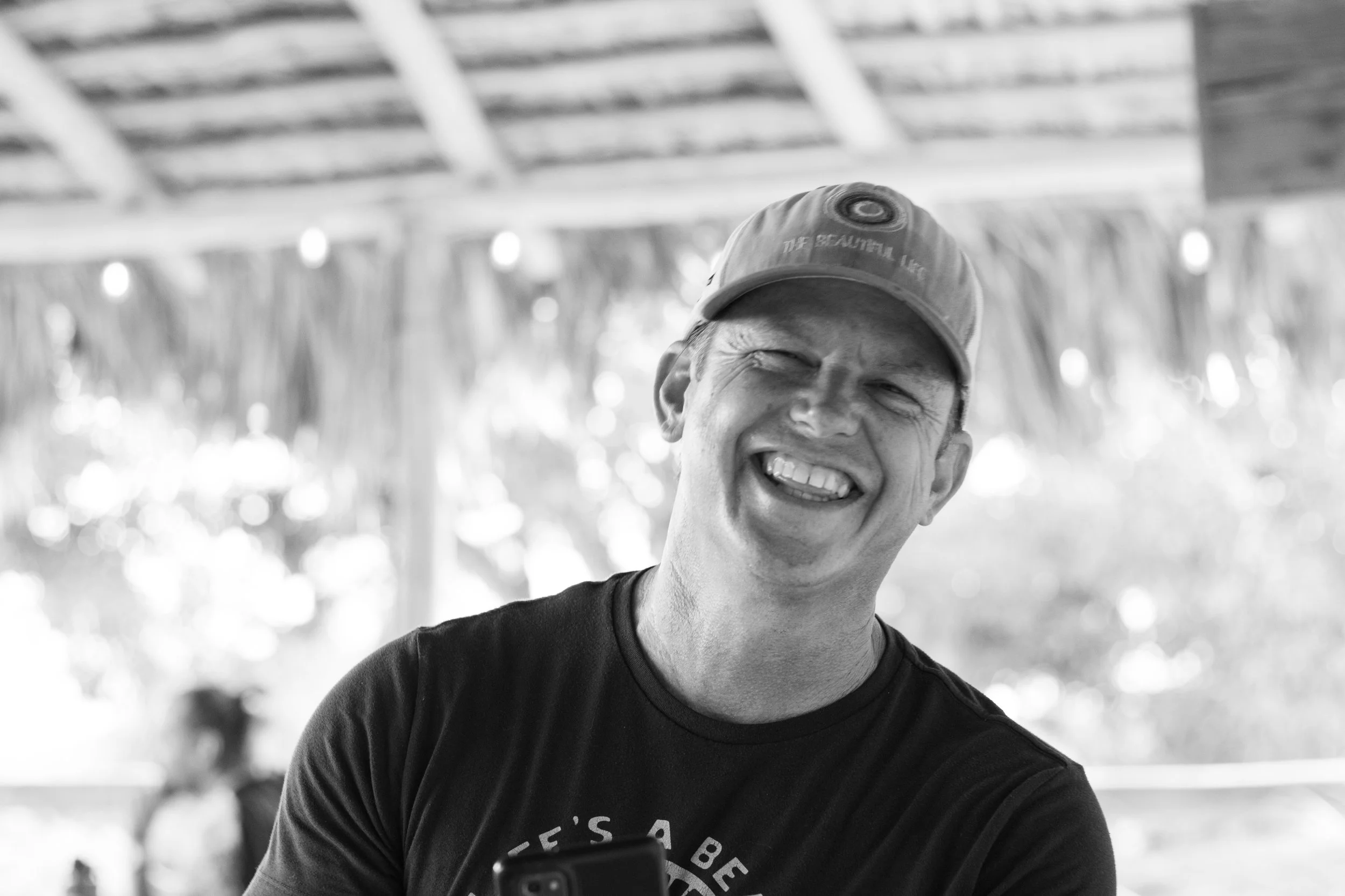 A man smiling and winking in a black and white photo, wearing a cap and t-shirt, with a blurry background of a thatched roof and trees.