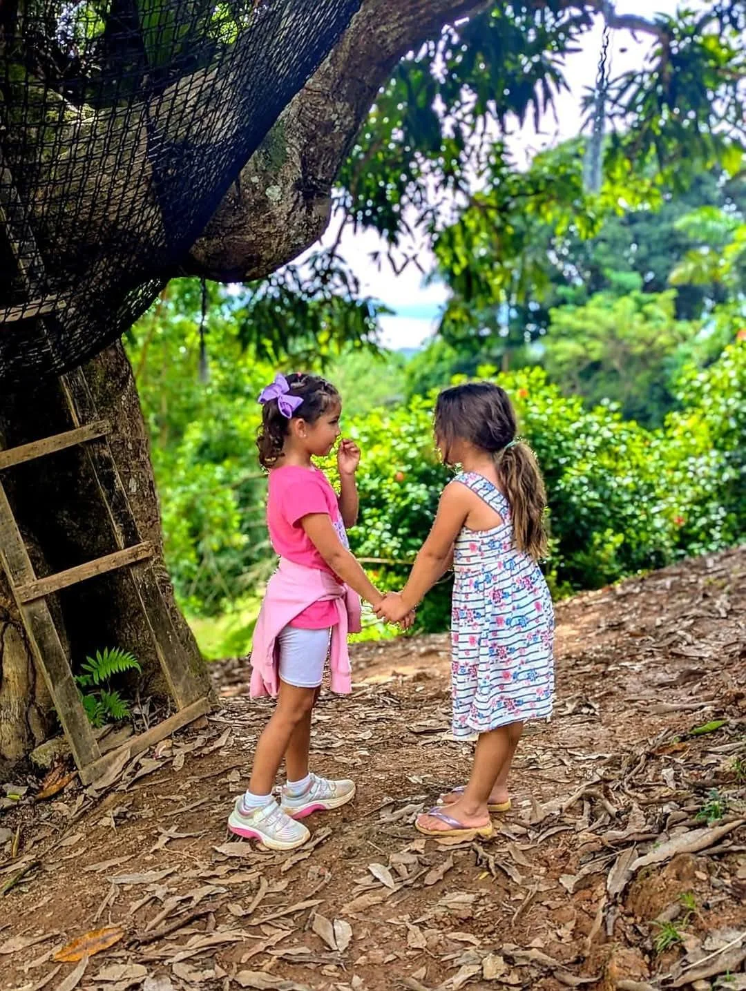 Two young girls holding hands and talking outdoors on a dirt path surrounded by green trees and bushes.