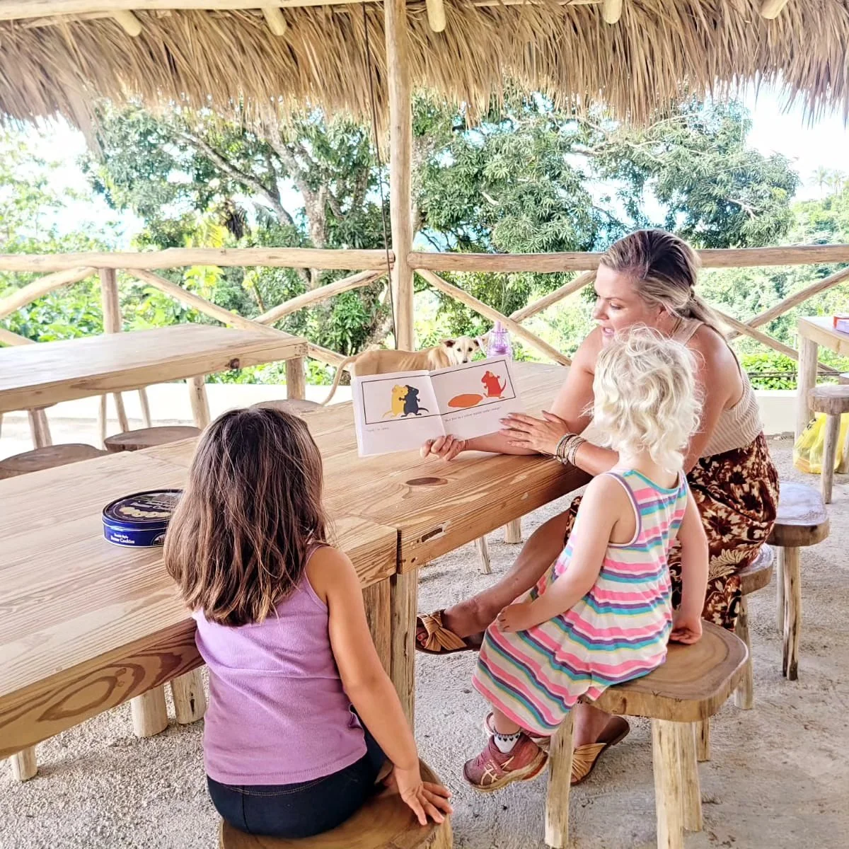 A woman reading a children's book to two young girls at an outdoor table under a thatched roof, with a dog in the background.