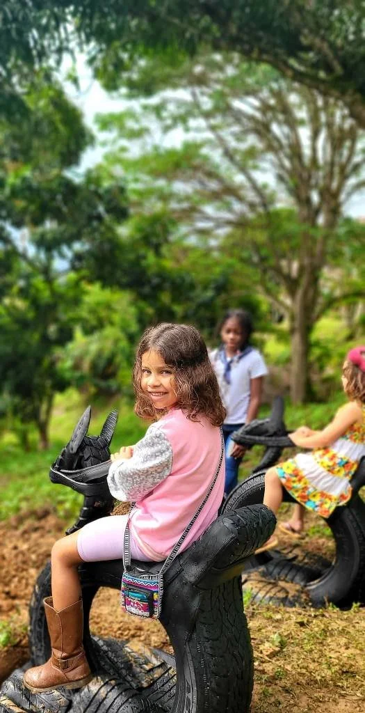 Children playing outdoors on tire toys shaped like motorcycles in a park with green trees.