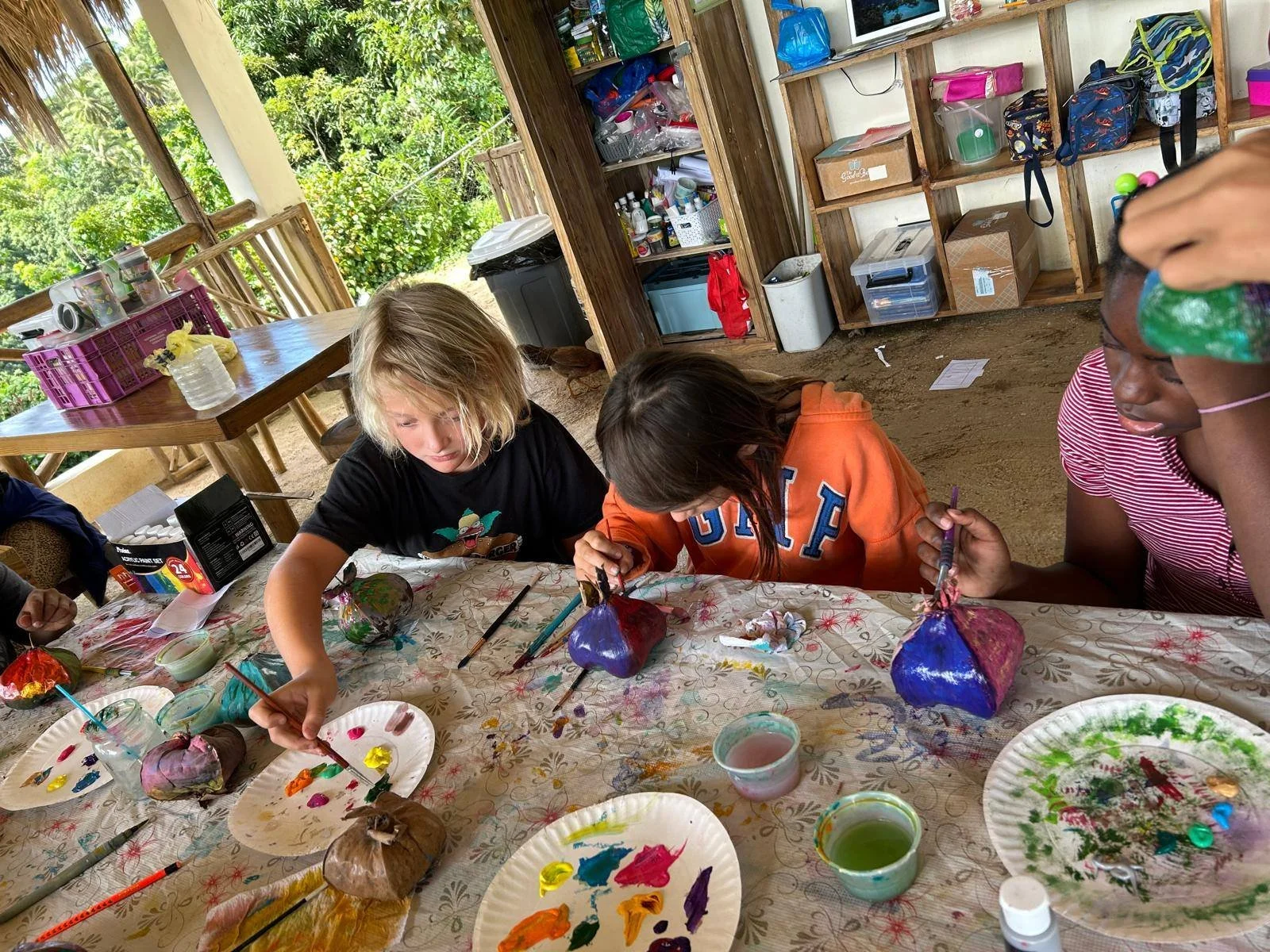 Children painting and decorating coconuts with colorful paints and brushes at a table in a rustic outdoor setting.
