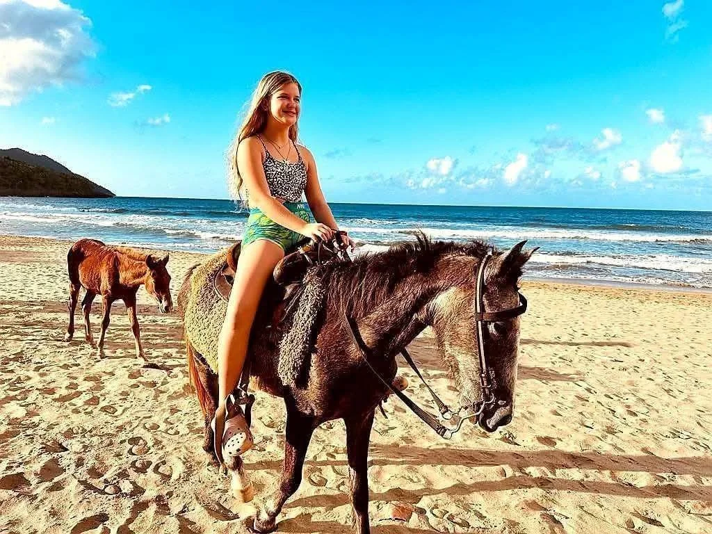 A girl riding a horse on the beach with two other horses nearby, ocean waves in the background, and a blue sky overhead.