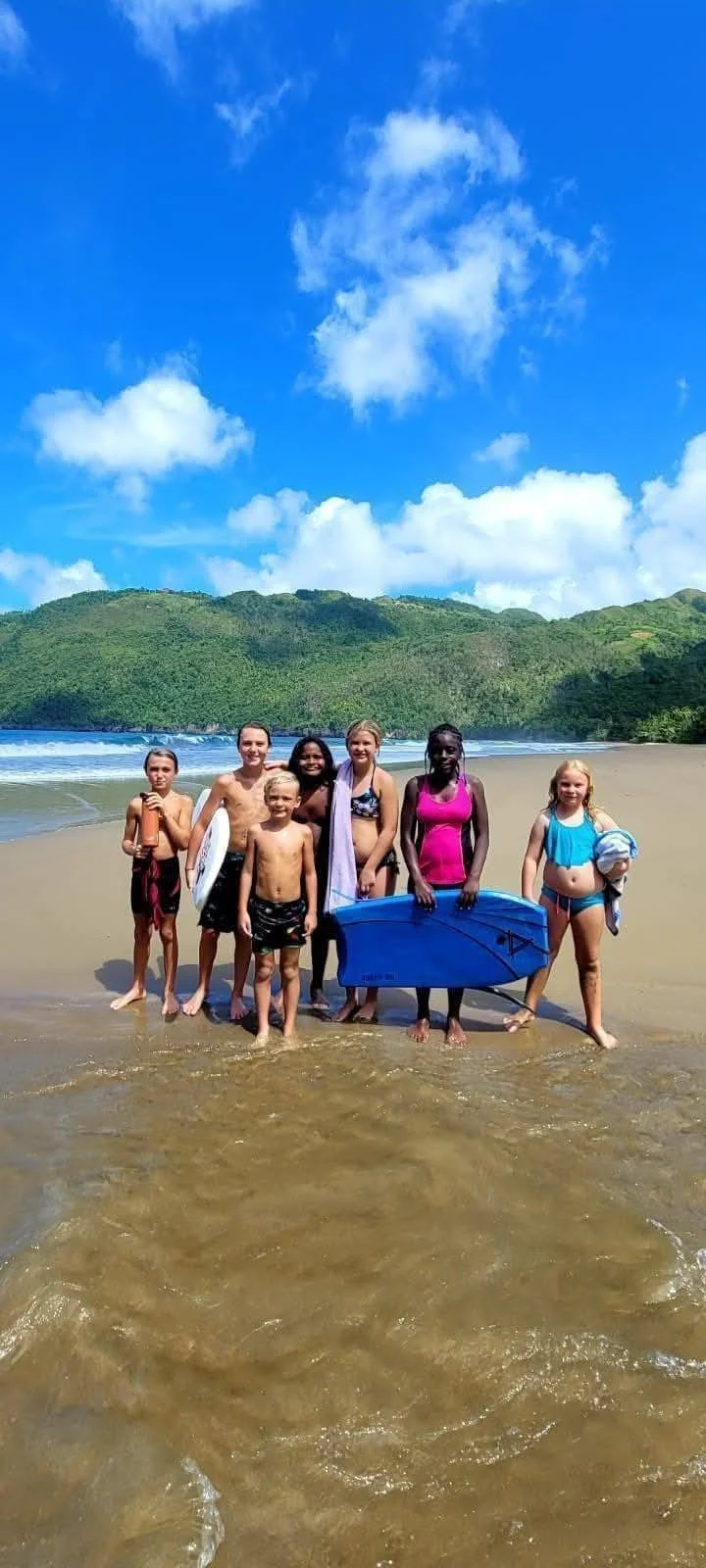 A group of children standing together on the beach, holding a surfboard, with a scenic background of green hills, blue sky, and white clouds.