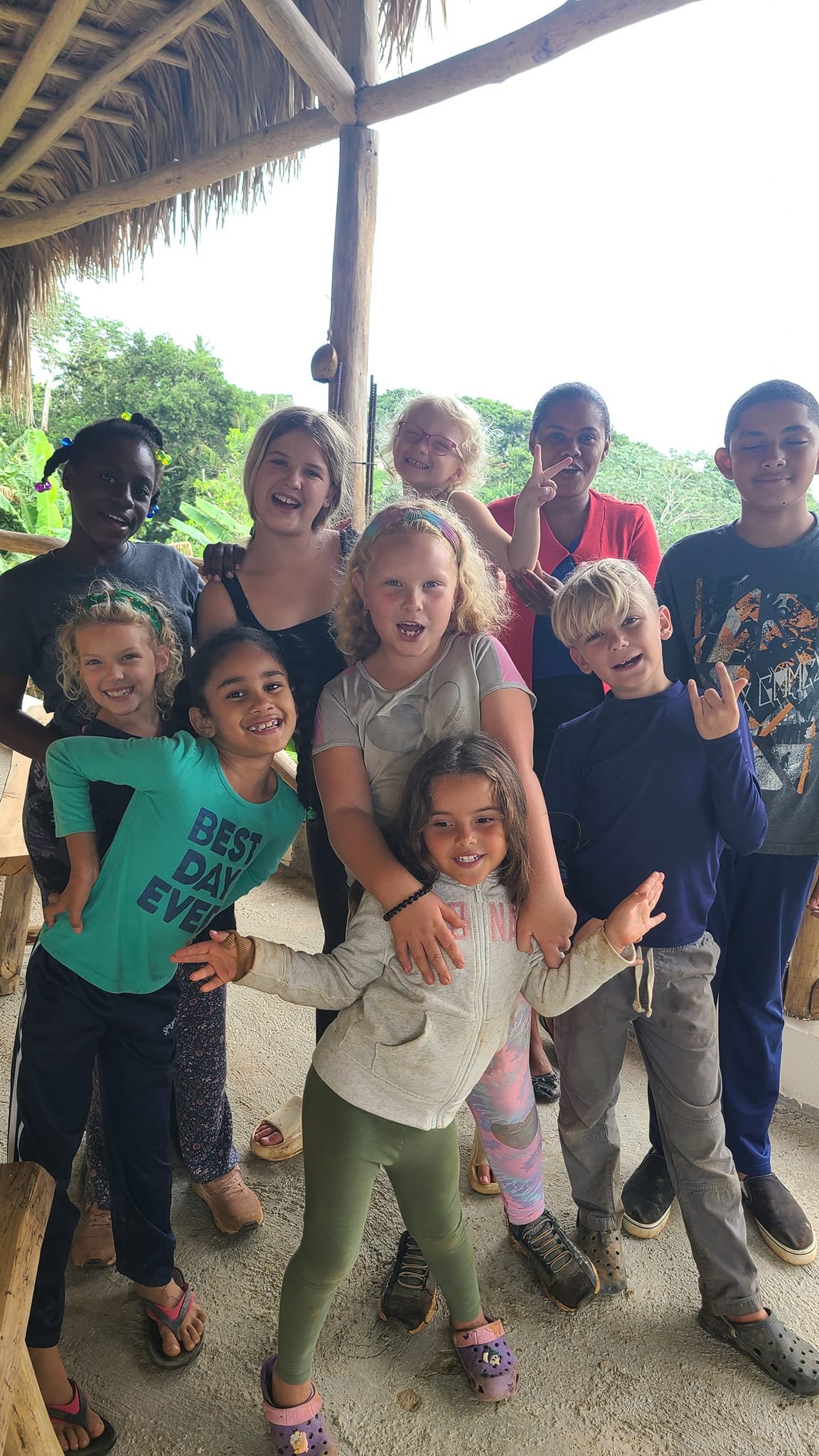 Group of children and a woman smiling and posing together outside under a thatched roof with green trees in the background.