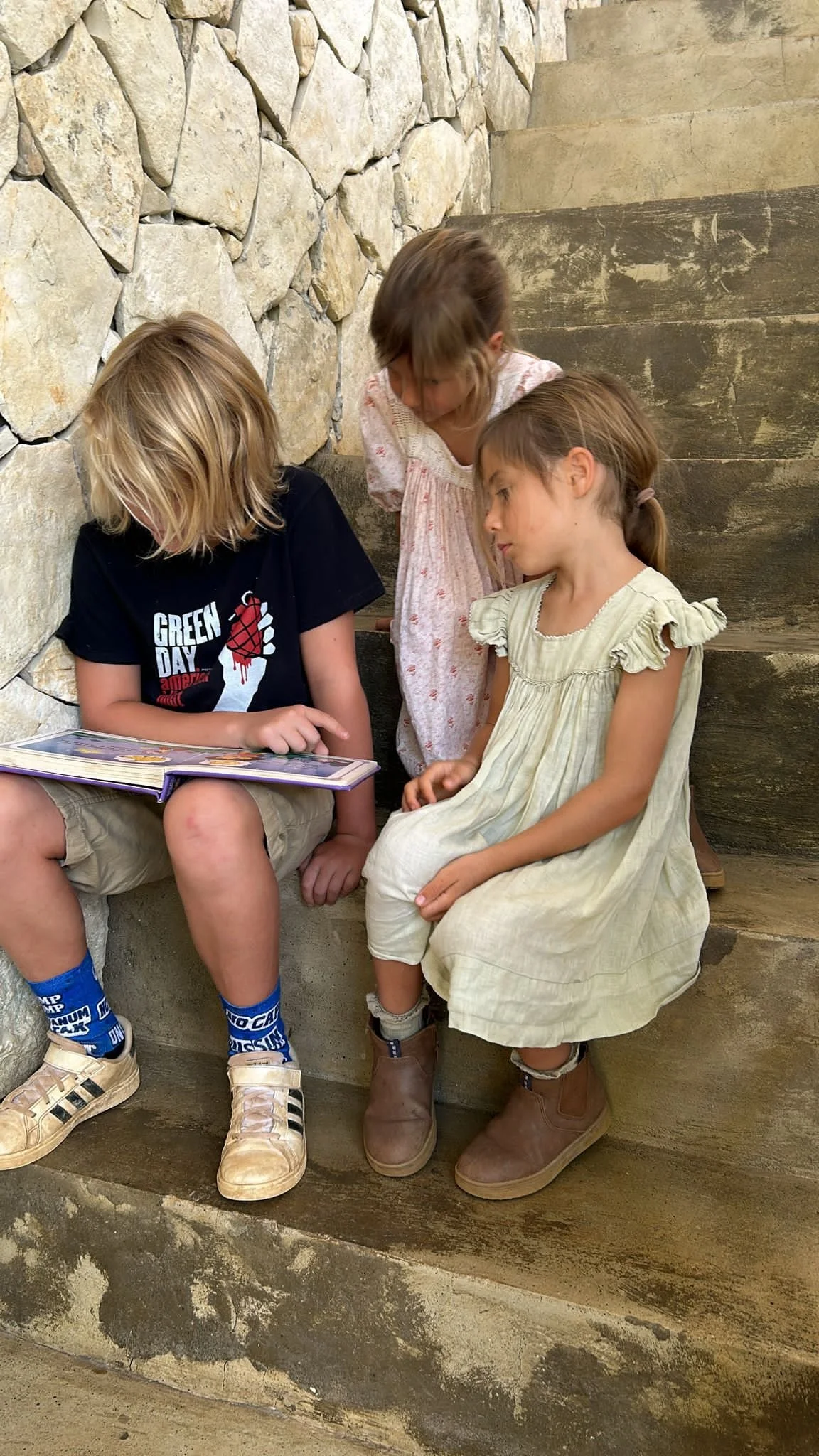 Three children sitting on stone and wooden steps, looking at a book.