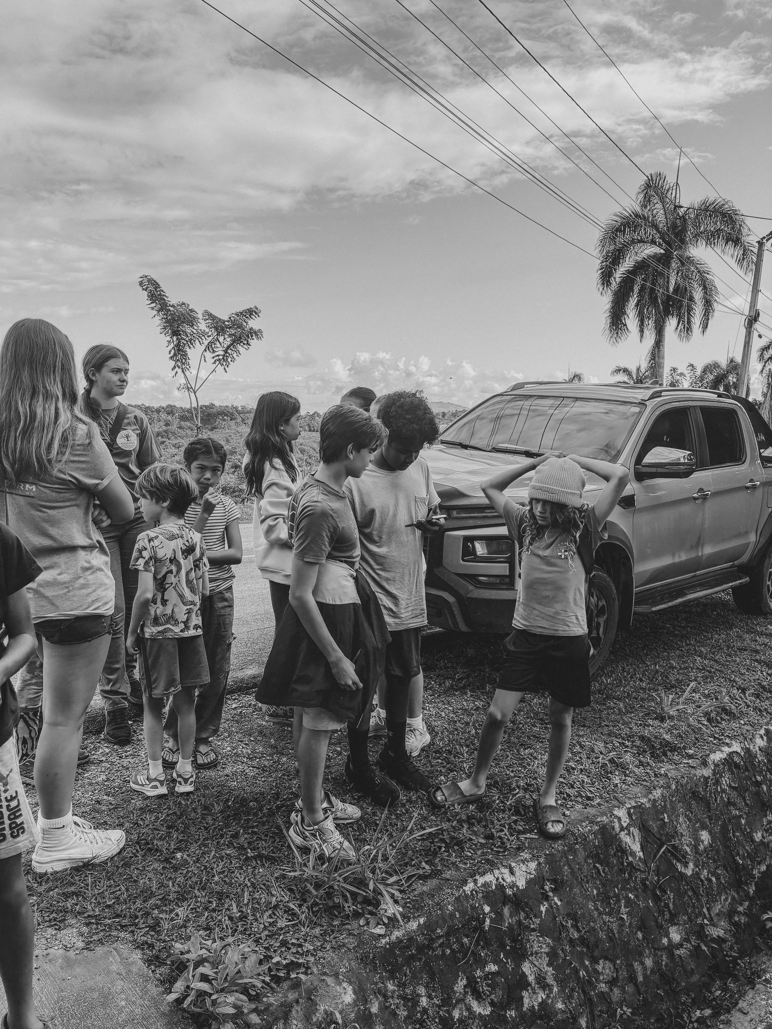 A group of children and teenagers gathered outdoors near a parked SUV, with some looking at a girl who is standing at the edge of a small ditch. There are palm trees and power lines in the background under a cloudy sky.