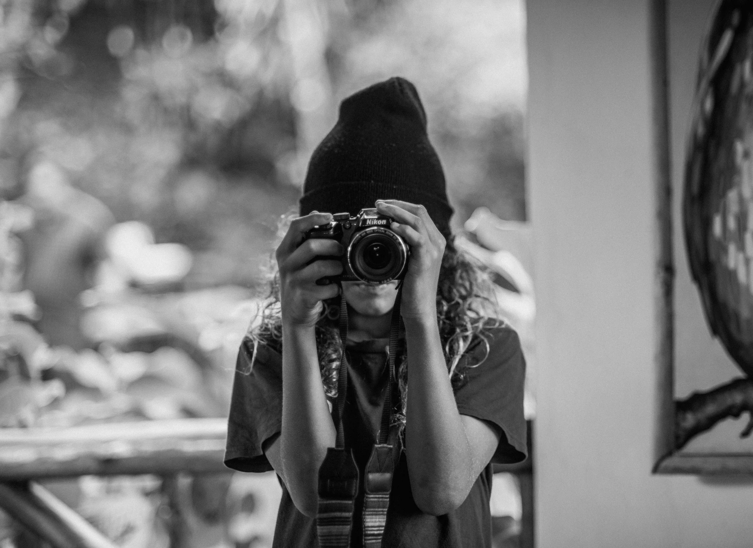 A person with curly hair wearing a beanie hat, holding a Nikon camera up to their face, taking a photo in black and white.