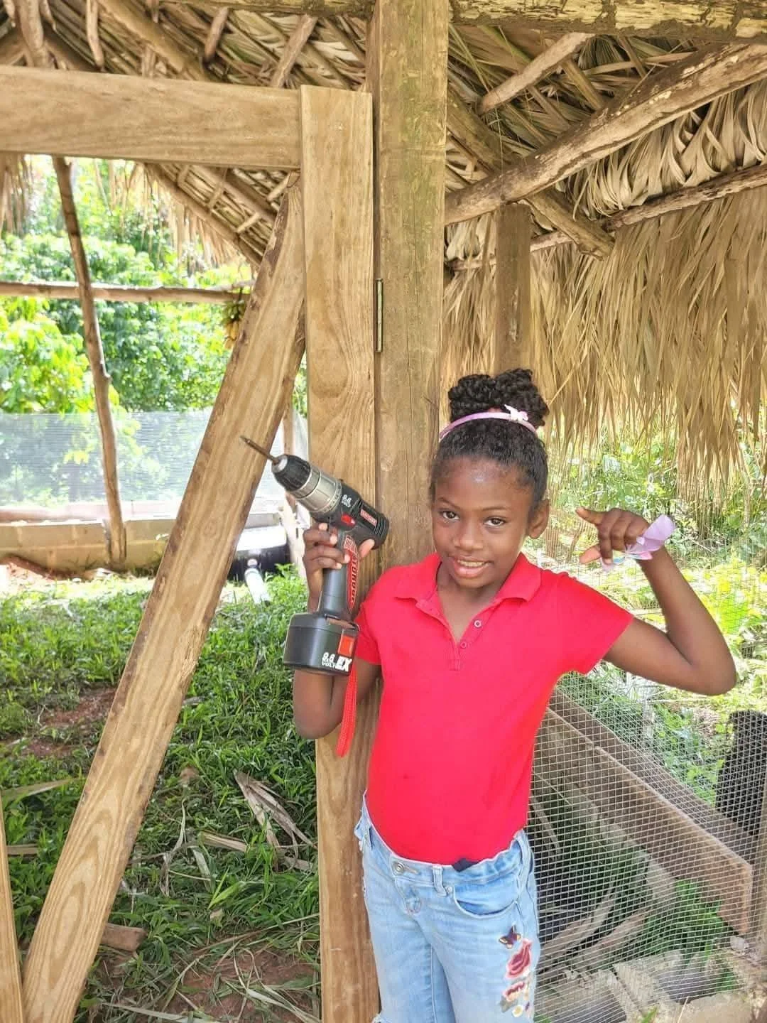 A young girl with braided hair, wearing a red polo shirt and light blue jeans, stands inside a wooden and thatched hut, holding a cordless drill in her right hand and smiling at the camera. The hut appears to be under construction with green plants a