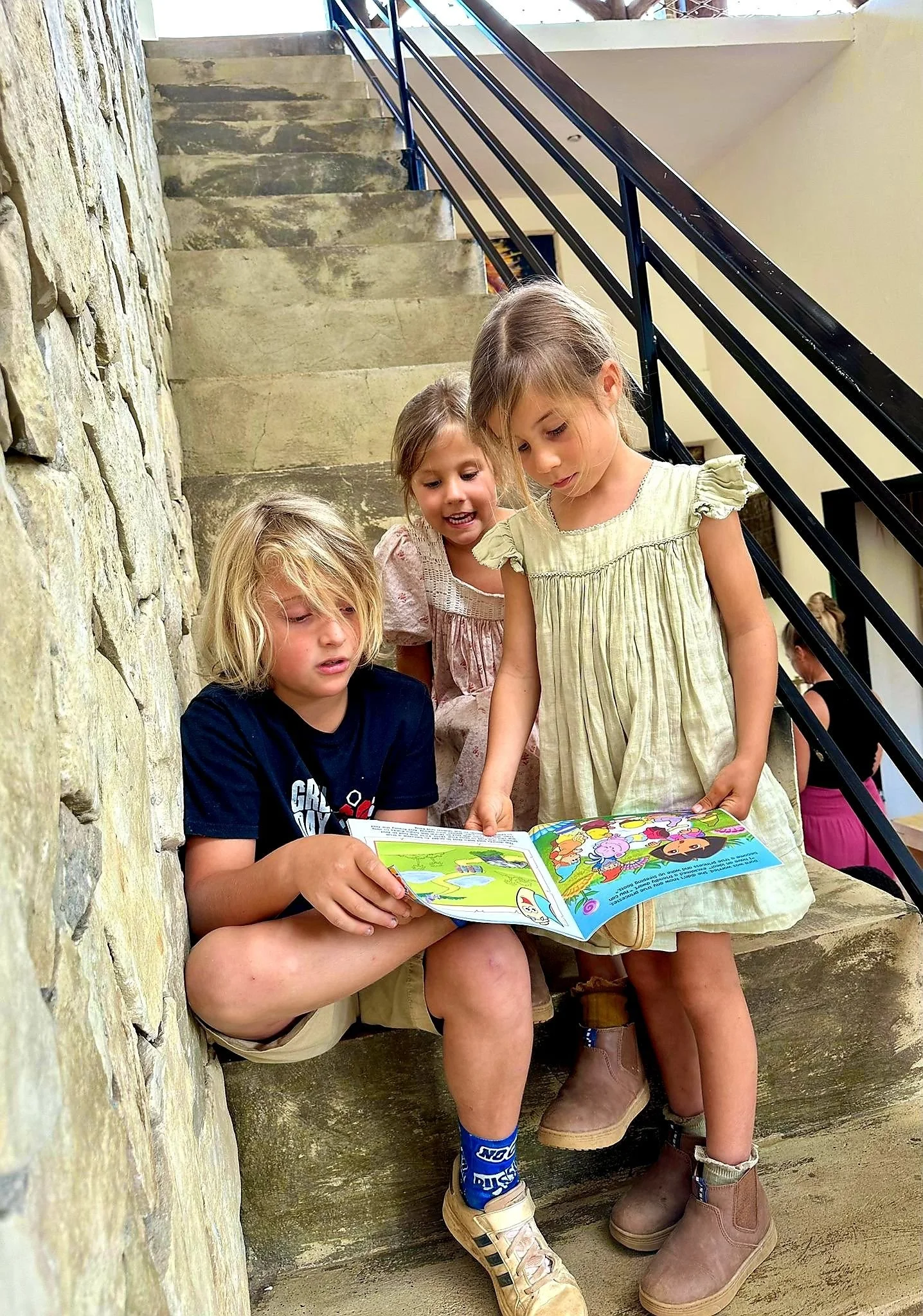 Three young children sitting on stone stairs, reading a colorful children's book, with a stone wall on one side and a metal railing on the other.