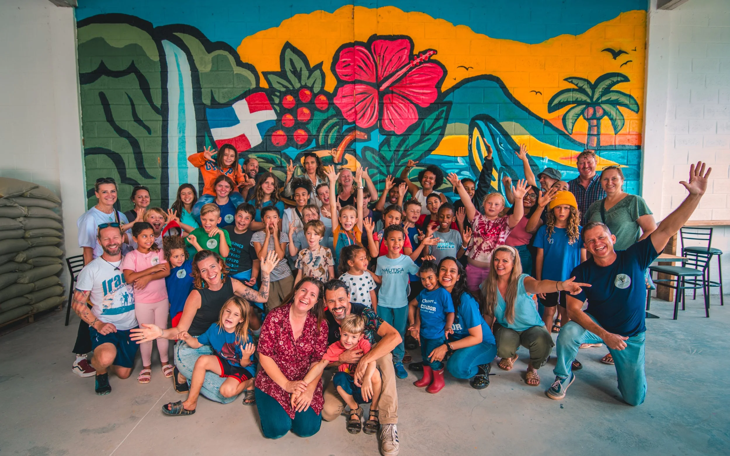 A large group of smiling children and adults posing in front of a colorful mural of tropical plants, volcanoes, and a sunset. The group includes boys and girls of various ages and a few adults, some of whom are kneeling or sitting in front, with ever