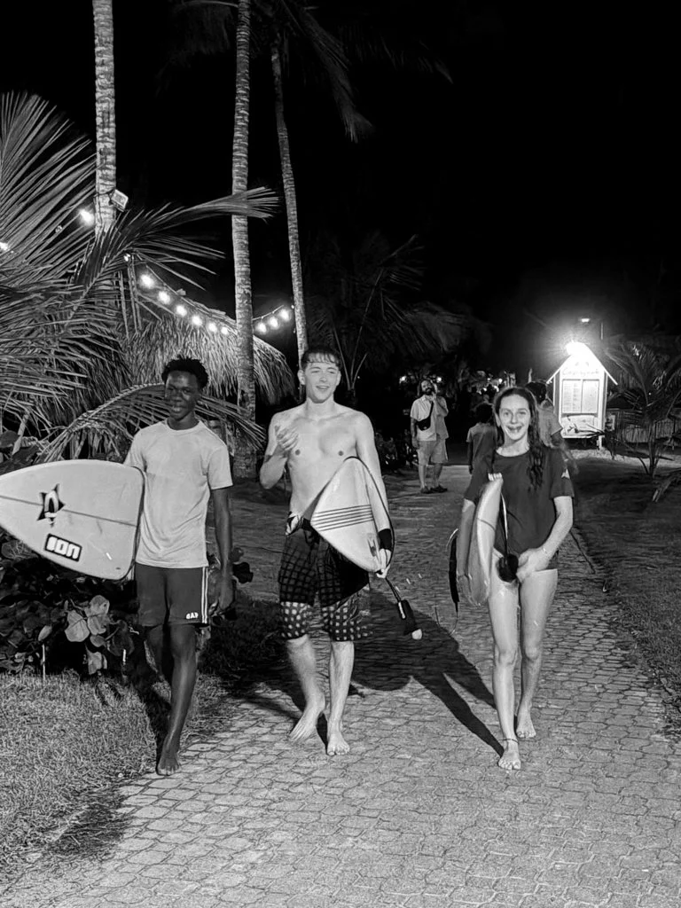 Three people walking on a cobblestone path at night, carrying surfboards, with palm trees and string lights in the background, in a tropical beach setting.