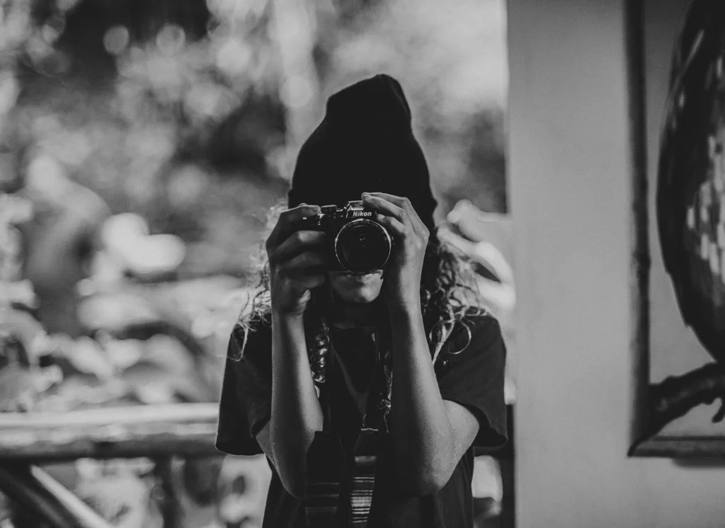 A person wearing a beanie taking a photo with a camera in a black-and-white setting.