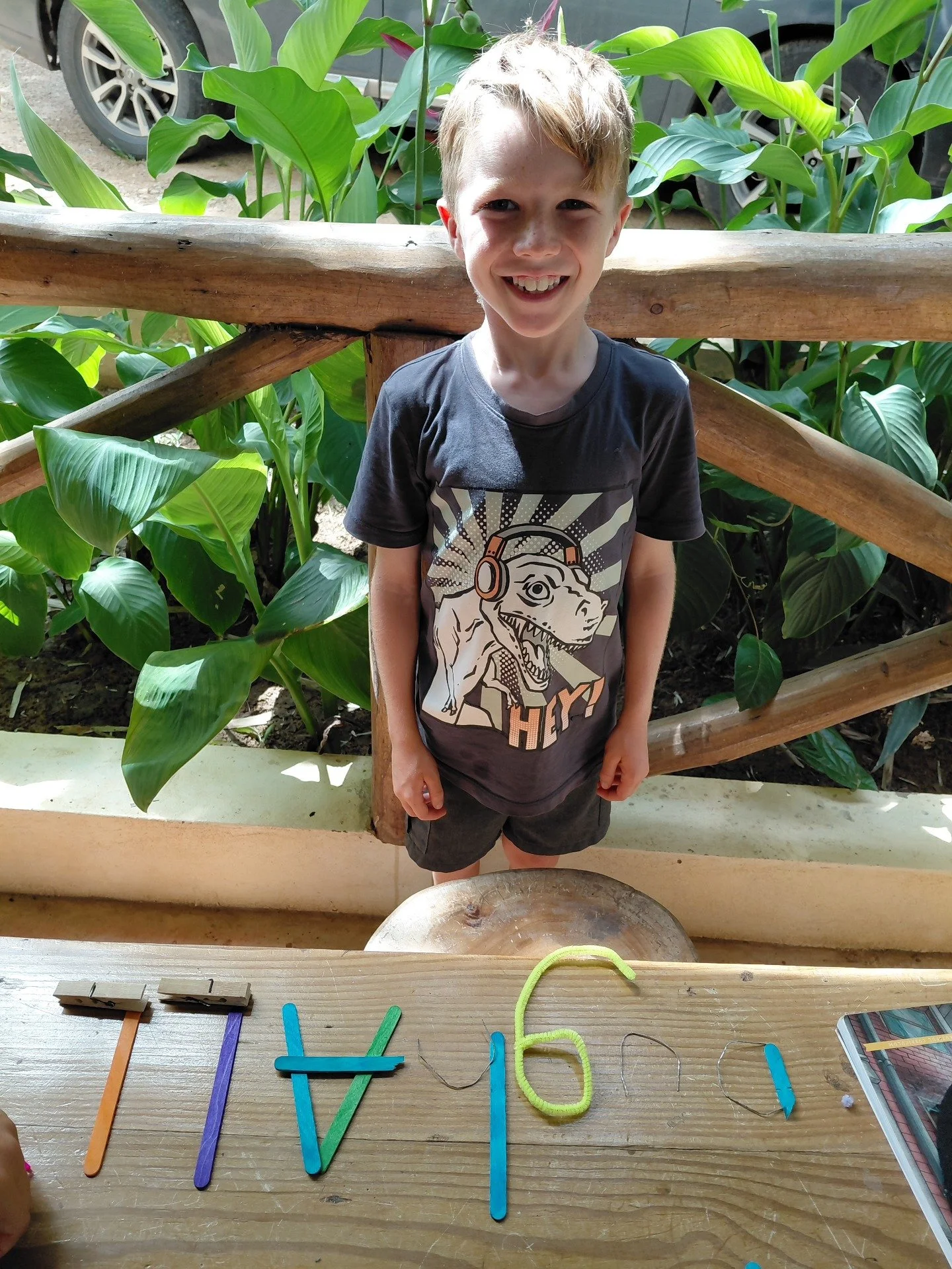 A young boy with short blond hair smiling, standing outdoors behind a wooden railing, surrounded by green tropical plants, wearing a black T-shirt with a cartoon dinosaur wearing headphones and the text 'HEY' on it, positioned behind a wooden table w