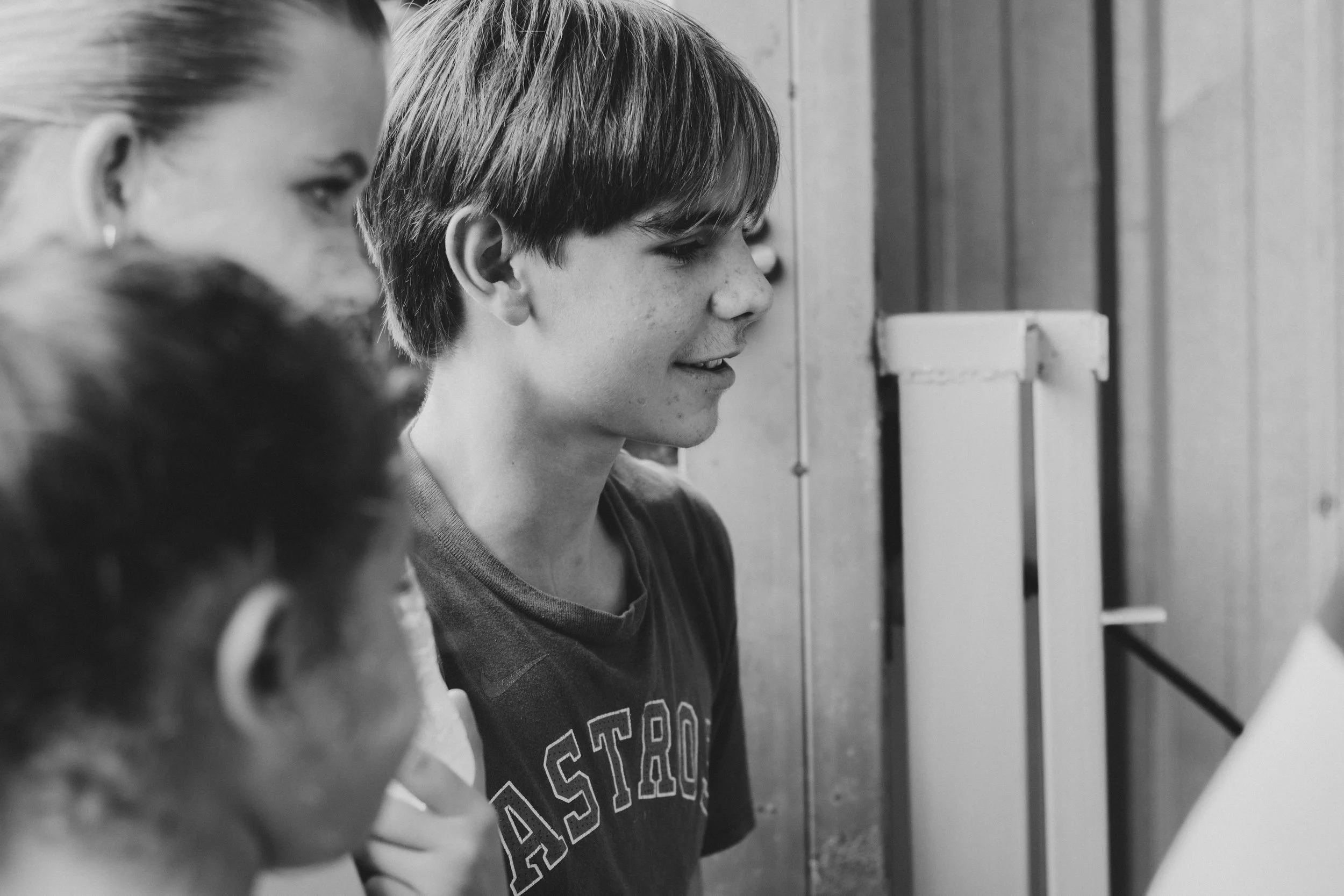 Three young people, two girls and one boy, are closely looking at a computer or screen, with the boy smiling. They are in an indoor setting with wood-paneled walls.