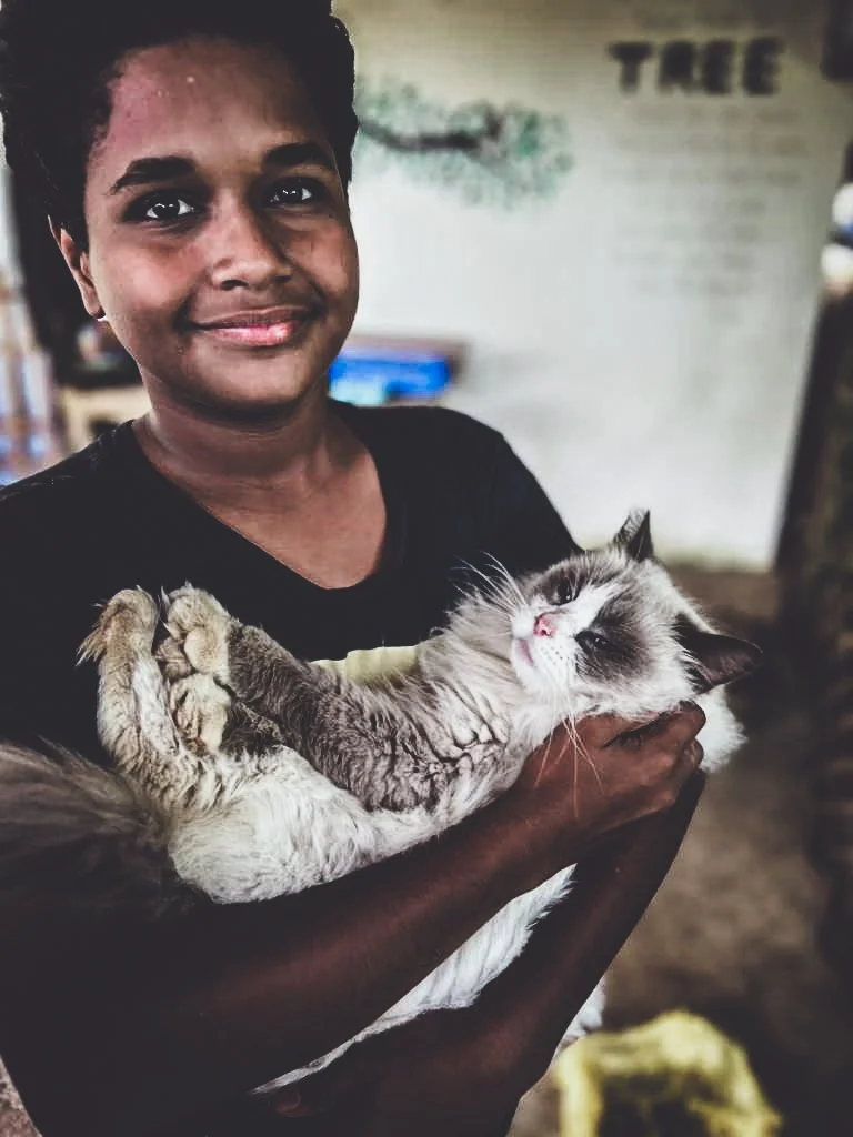 A young person with dark skin and short curly hair smiling while holding a relaxed Siamese cat, indoors with blurred background.