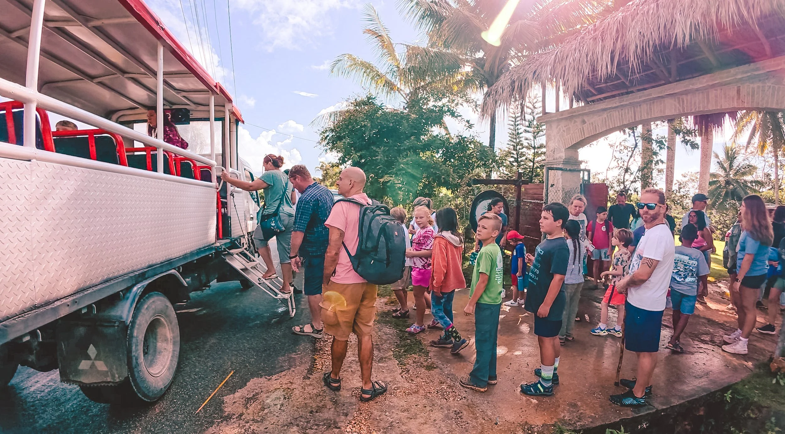 Group of people, including children and adults, boarding a double-decker bus in a tropical setting with palm trees and a thatched-roof structure in the background.