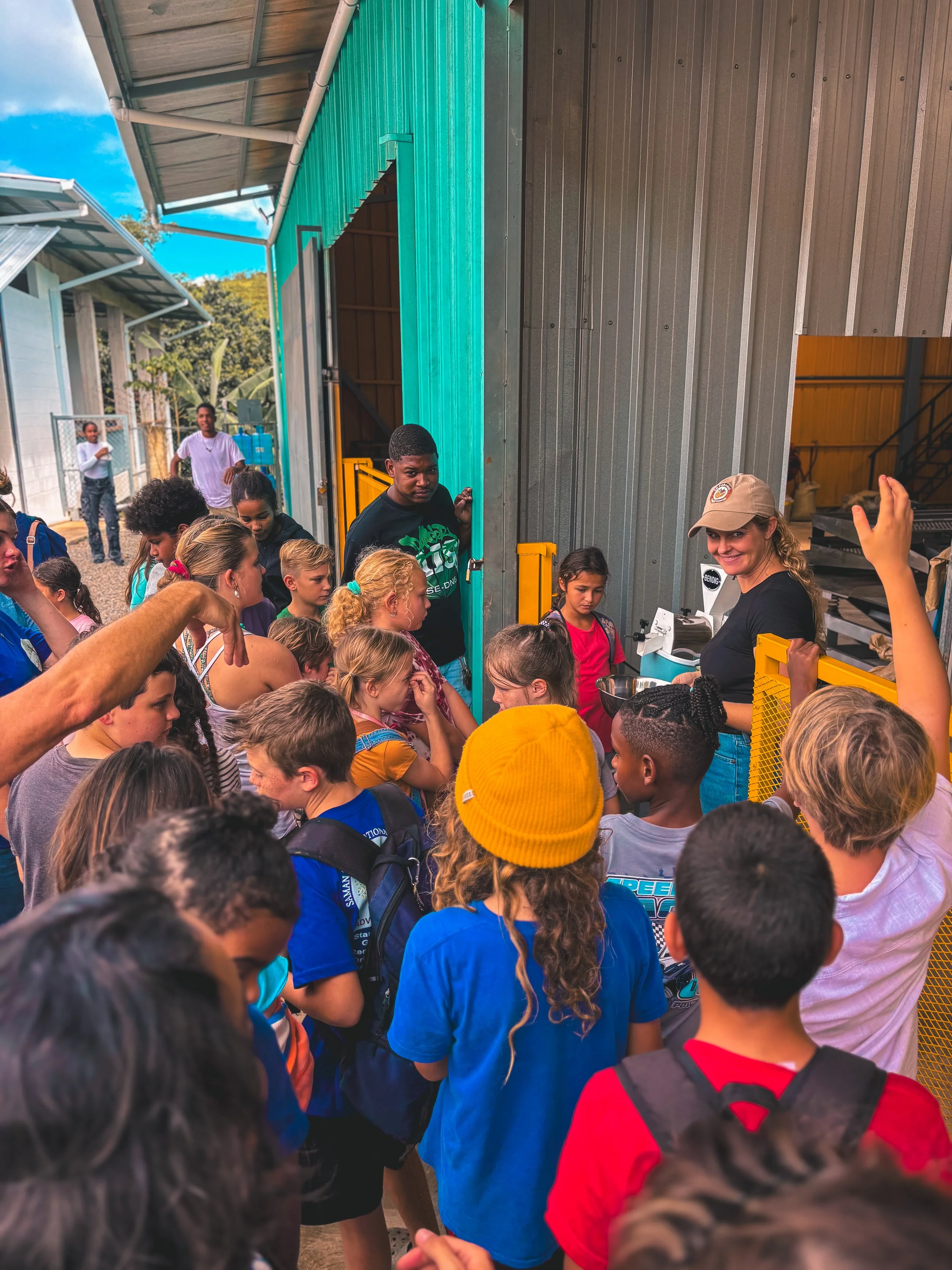 Group of children and a few adults gathered around a woman with a hat at an entrance to a building, likely listening to her during a trip or educational tour.