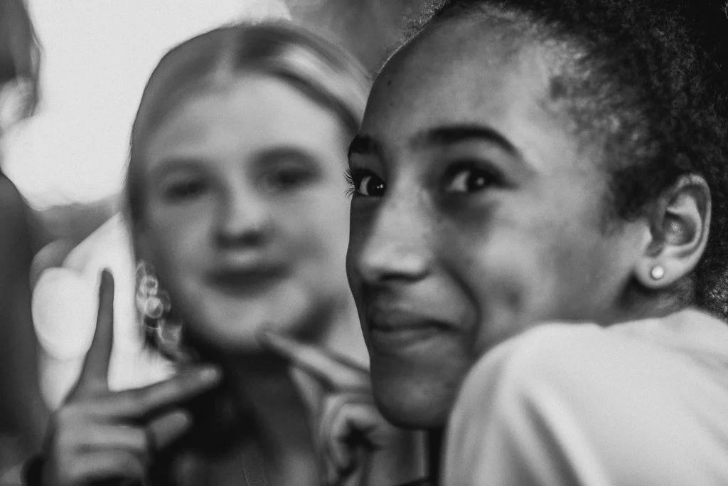 A close-up of a smiling young woman with curly hair and earrings, with another woman in the background making a peace sign.