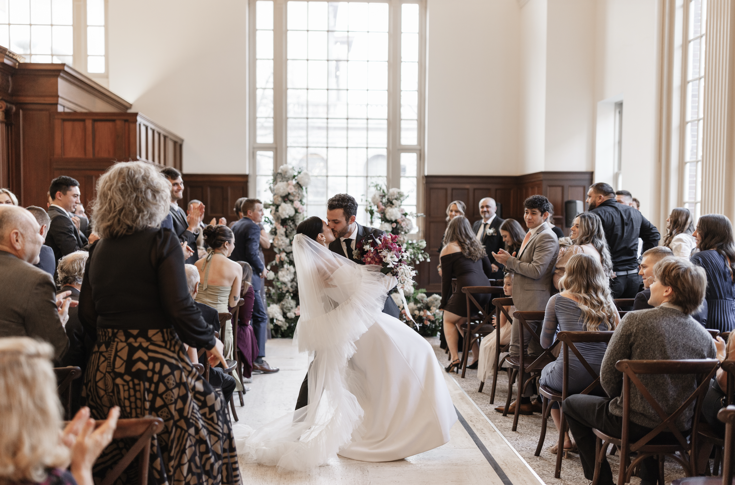 A bride and groom kiss during their wedding ceremony in a bright, spacious room with large windows and floral decorations, surrounded by seated guests.
