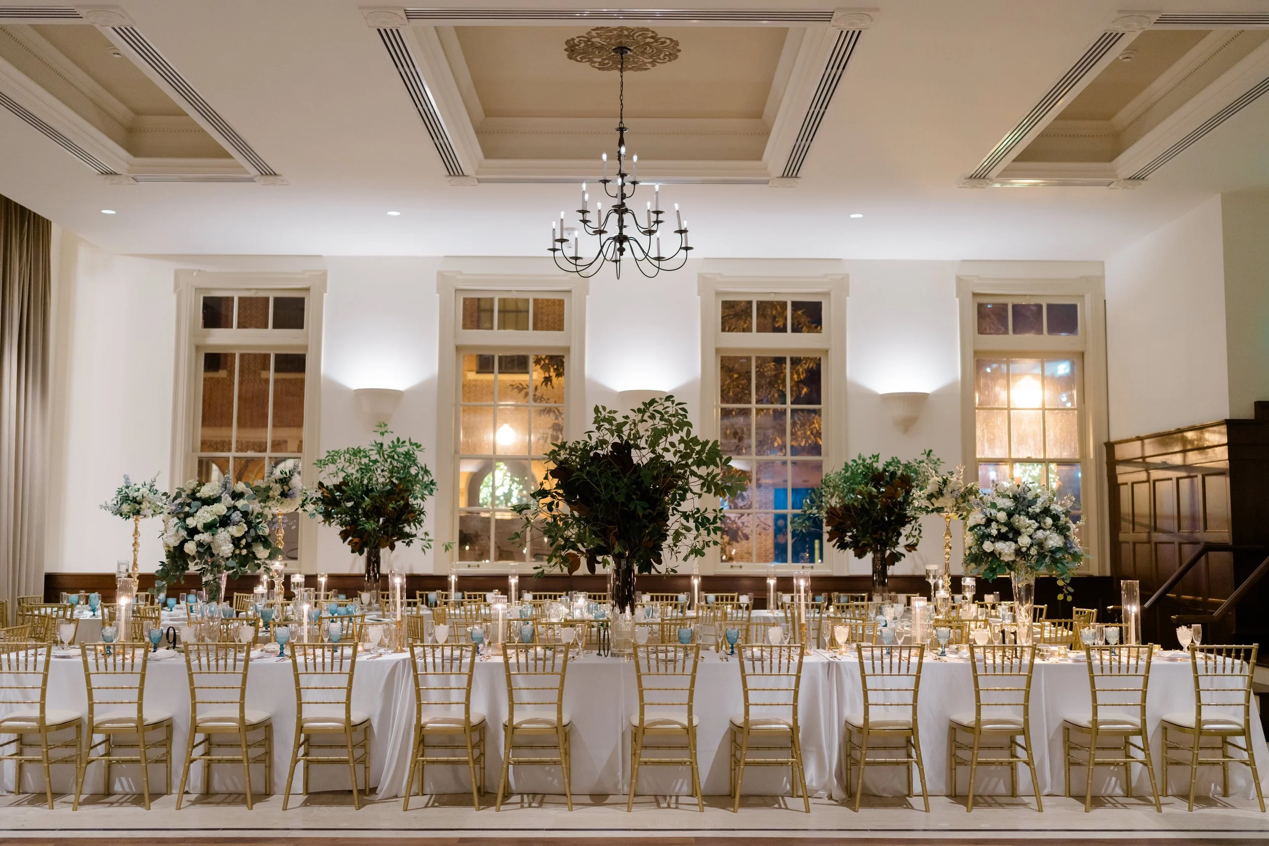 Elegant banquet hall with long head table draped in white tablecloth, set with glassware, candles, and floral centerpieces; gold chairs arranged around the tables; large floral arrangements and trees decoratively placed in the middle of the room, wit