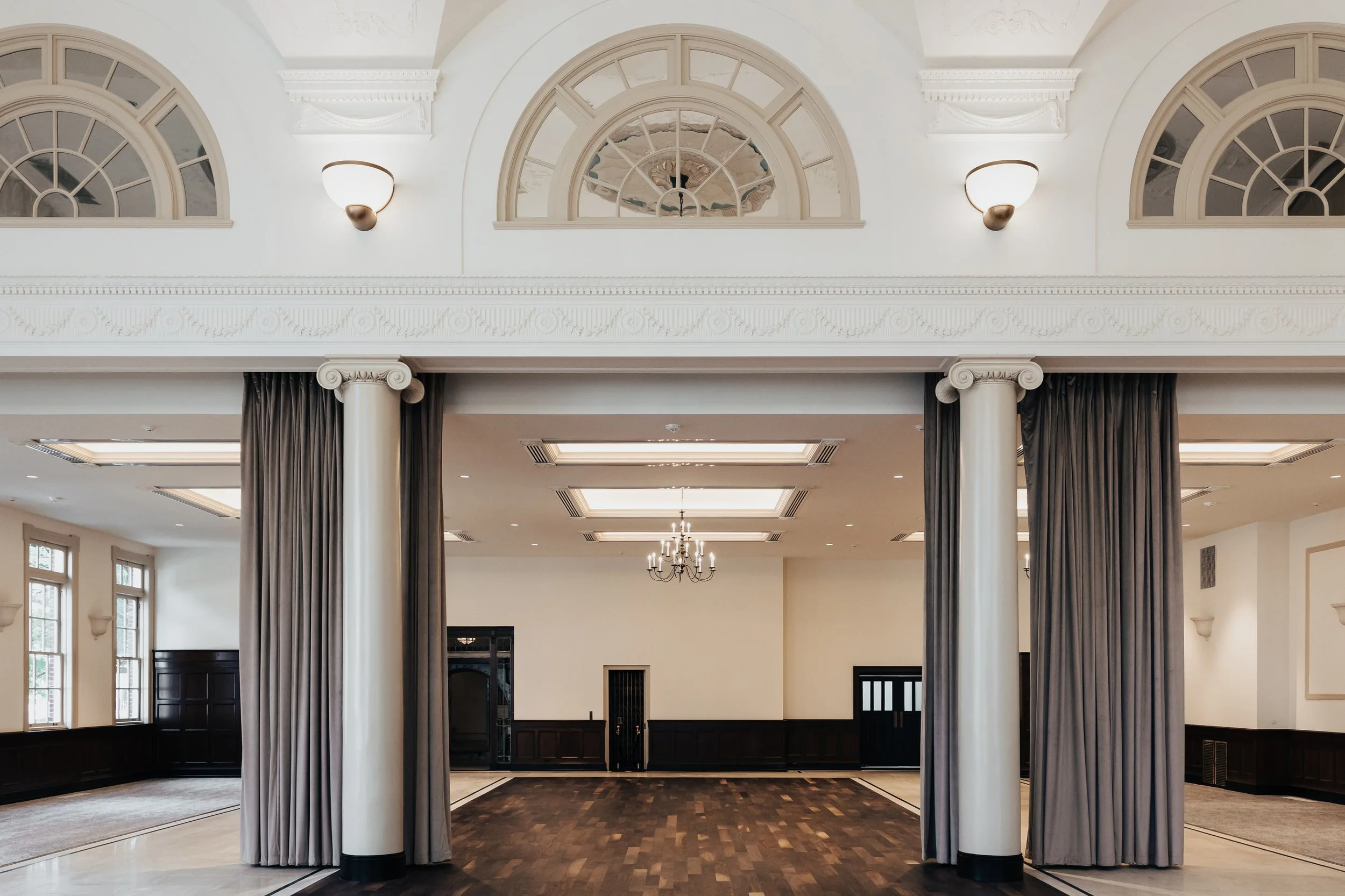 Interior of an elegant, spacious room with white walls, dark wood wainscoting, large windows, and a central open space with a chandelier, flanked by dark curtains and white columns.