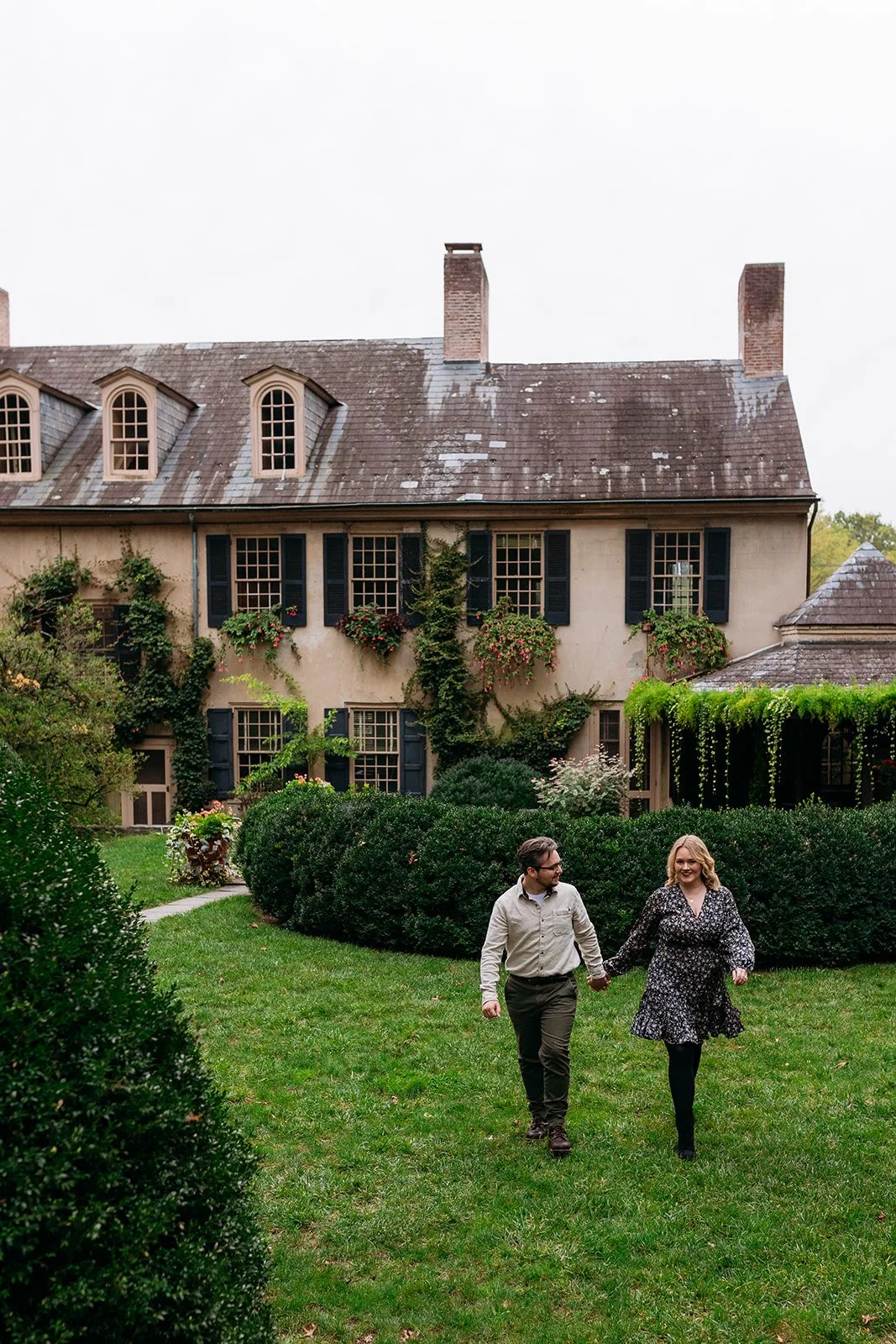 A man and woman holding hands walking on a green lawn in front of a large, old house with ivy and flower boxes.