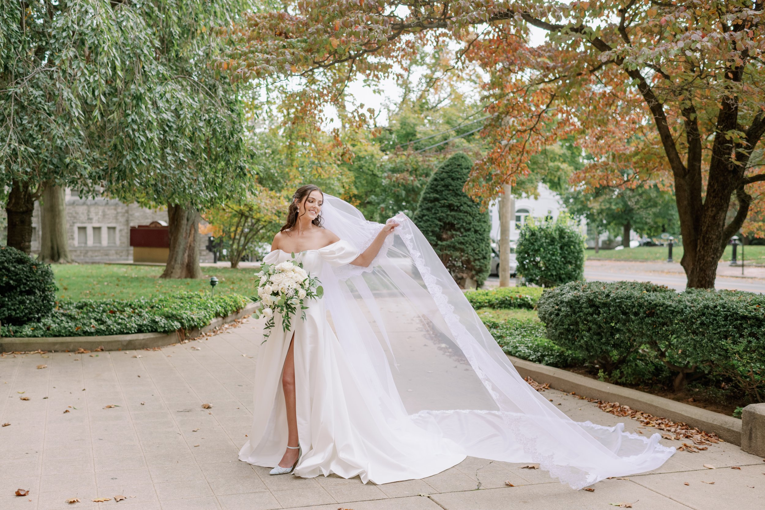 Bride in a white wedding gown holding a bouquet and lifting her veil in an outdoor garden setting during daytime, surrounded by trees with fall foliage.
