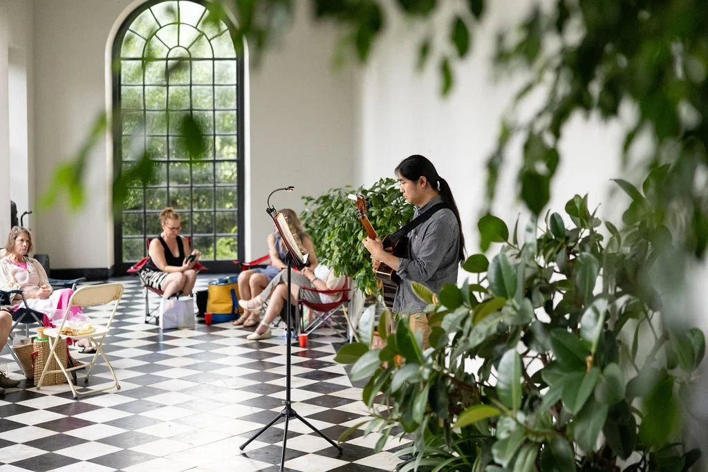 Woman playing guitar while three girls sit nearby in a sunlit room with tall arched window and checkered floor, surrounded by green plants.