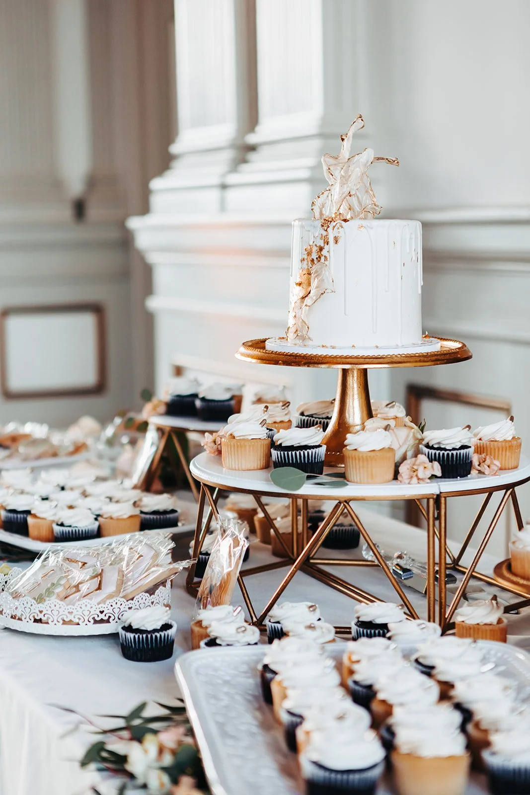 Elegant dessert table with a white tiered cake topped with dried flowers and surrounded by cupcakes and treats, set in a decorated room with large windows.