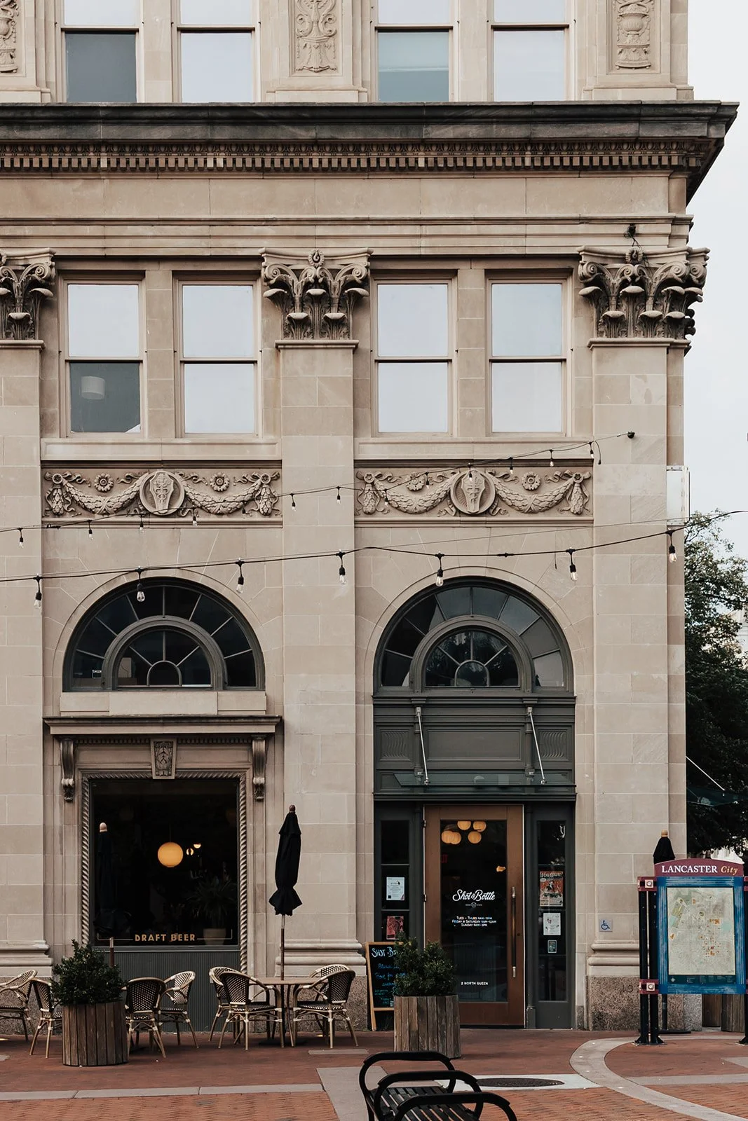 Front view of a historic building with ornate architectural details, two arched entrance doors, outdoor seating with tables and chairs, and string lights overhead.