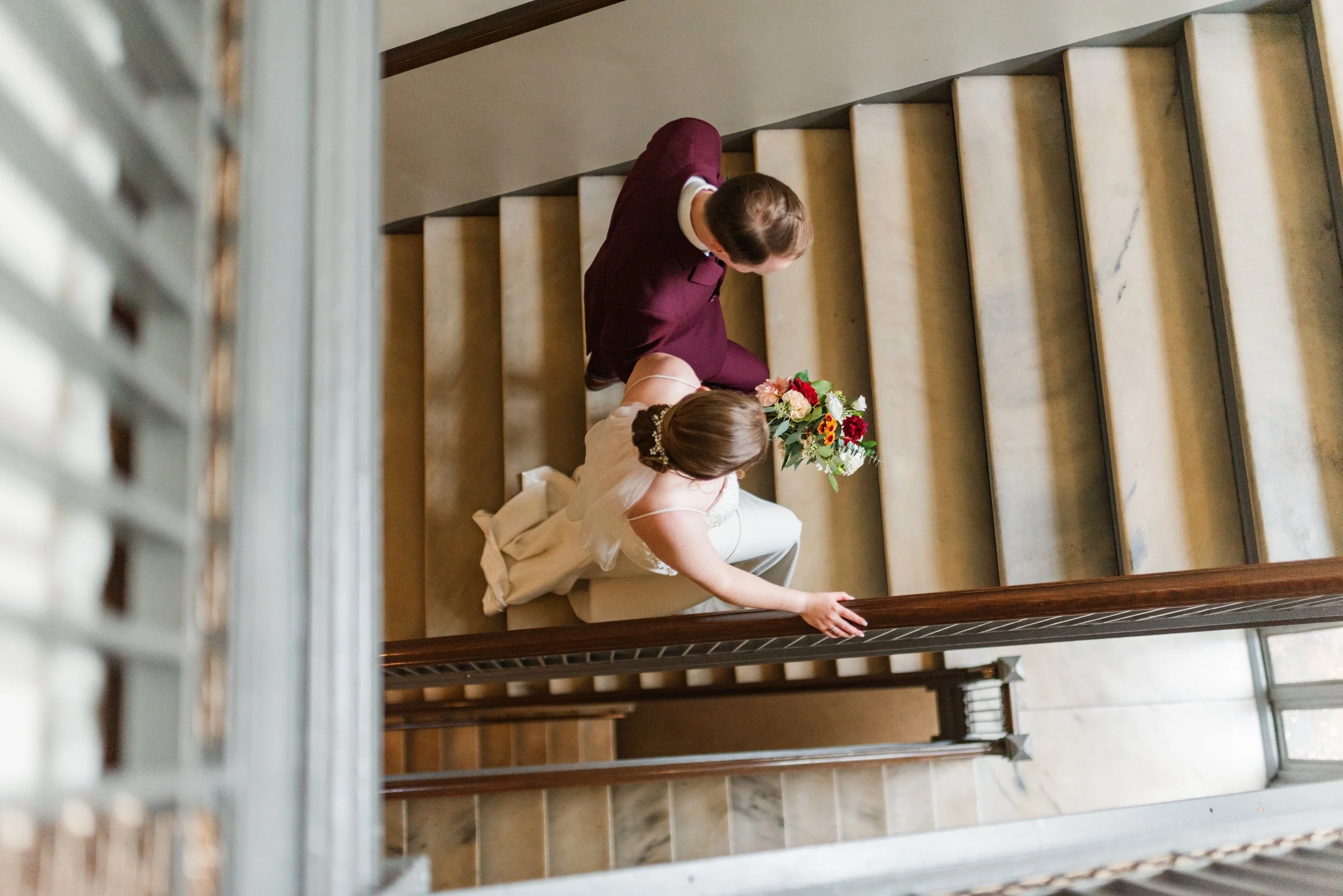 A bride in a white wedding dress holding a bouquet of flowers, walking down a staircase with a man in a burgundy suit, seen from above.